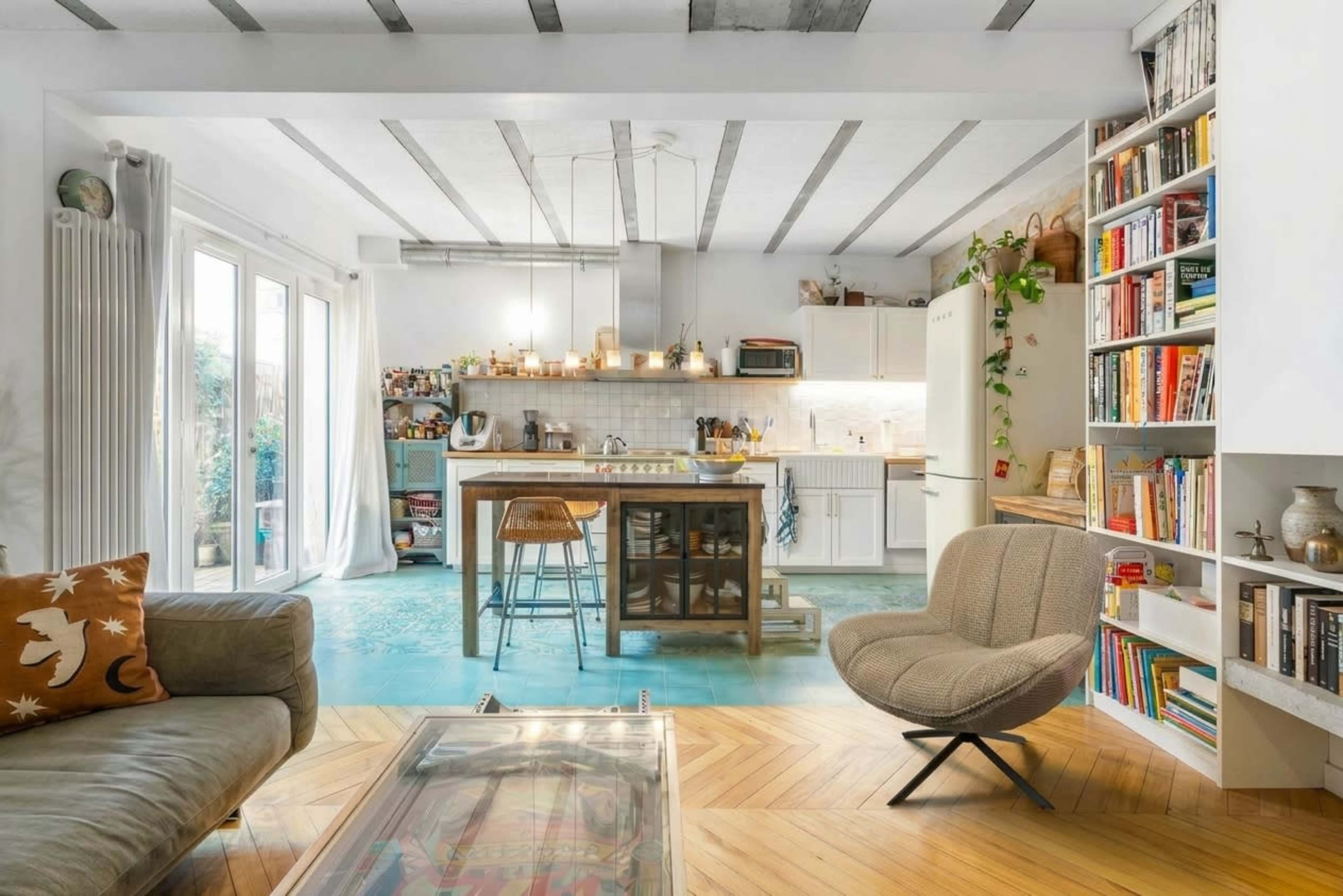 The image shows a modern kitchen and living area with a mix of cabinetry, a central island, and bookshelves displaying various items.