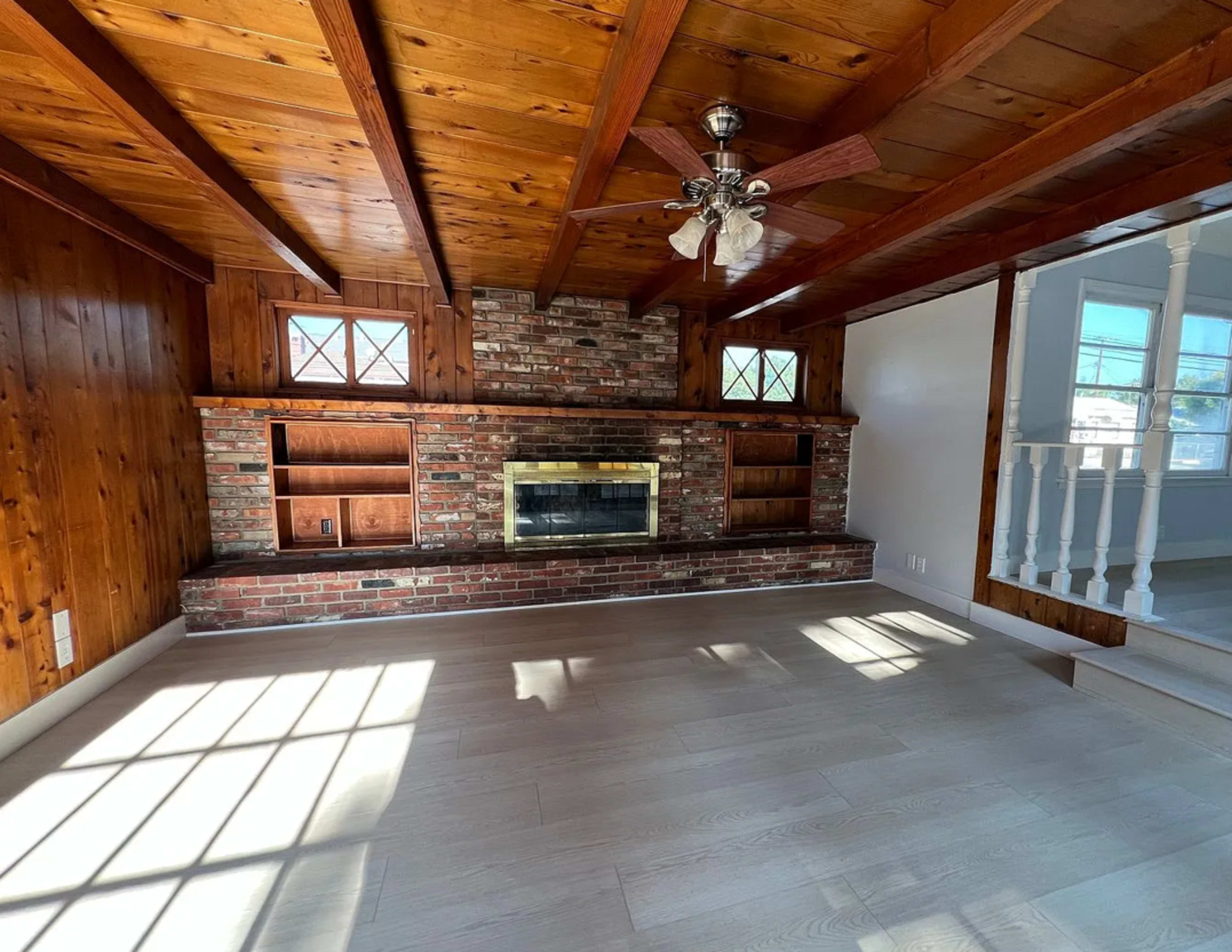 The image shows a living room with wooden beams on the ceiling, a brick fireplace, built-in shelves, and natural light streaming in through the windows.