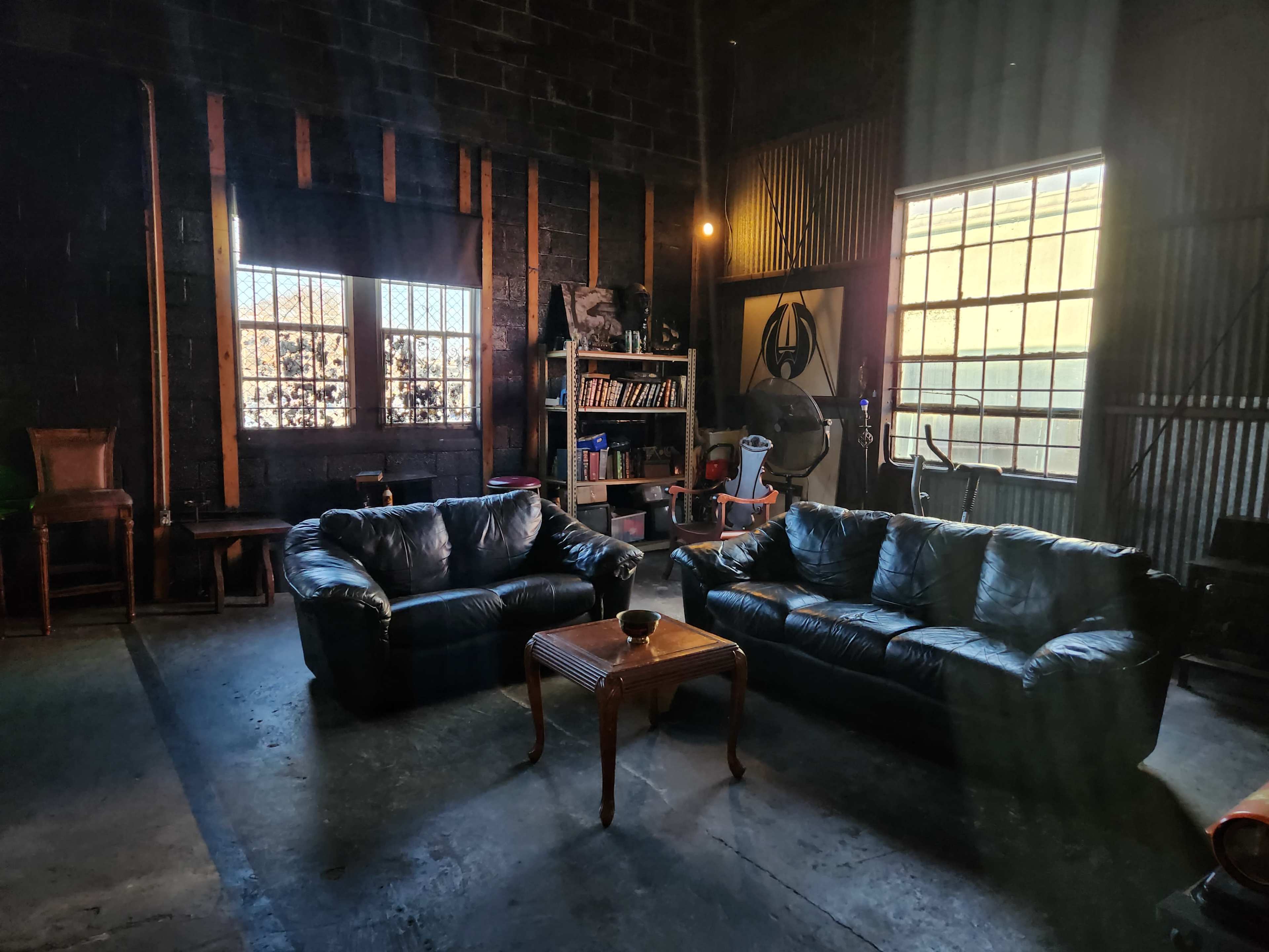 A dimly lit warehouse interior featuring two black leather sofas and a wooden coffee table, with large windows and various items visible in the background.