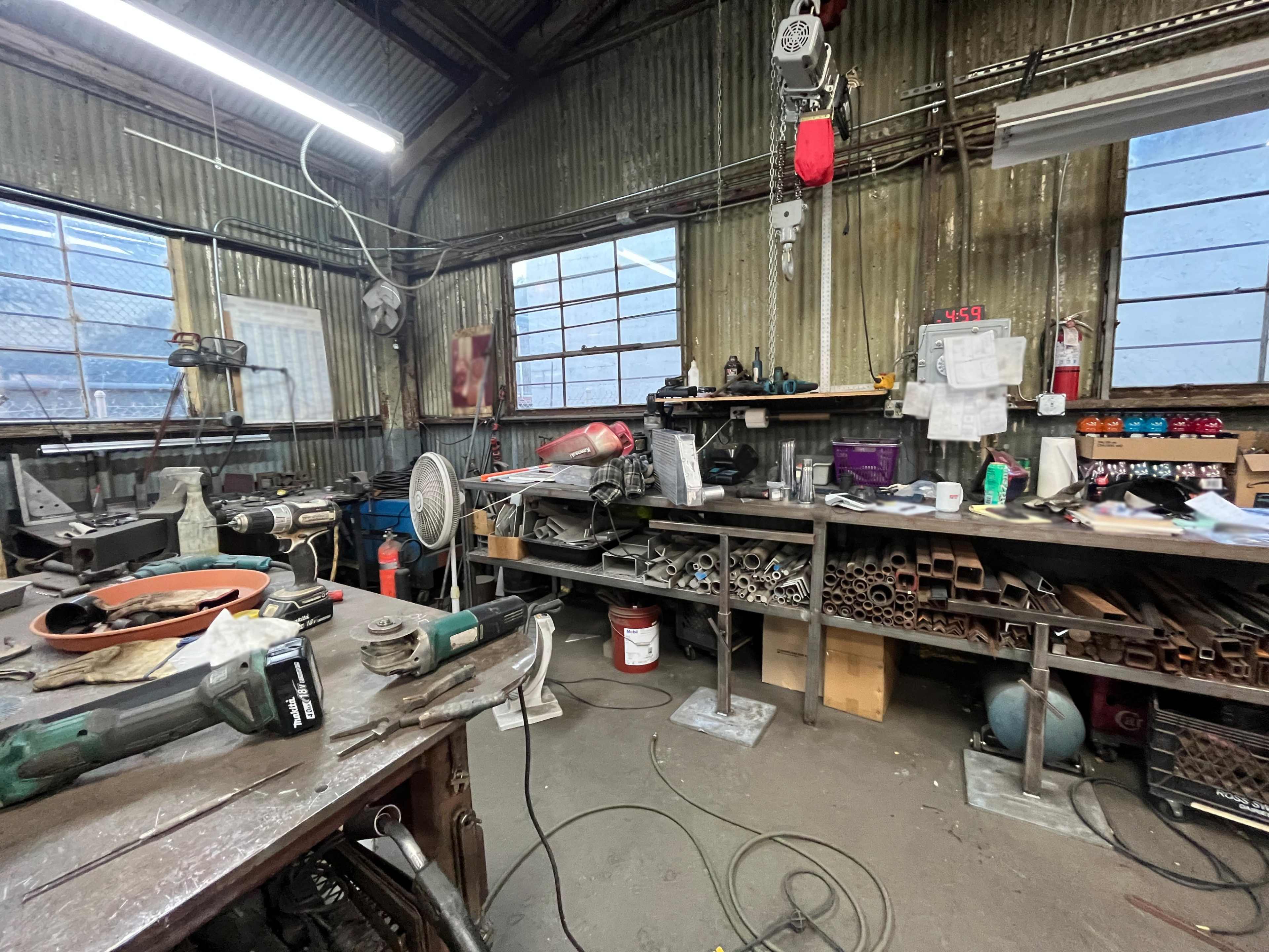 The image shows a cluttered workshop with various tools, metal pipes, and equipment scattered on workbenches and the floor.