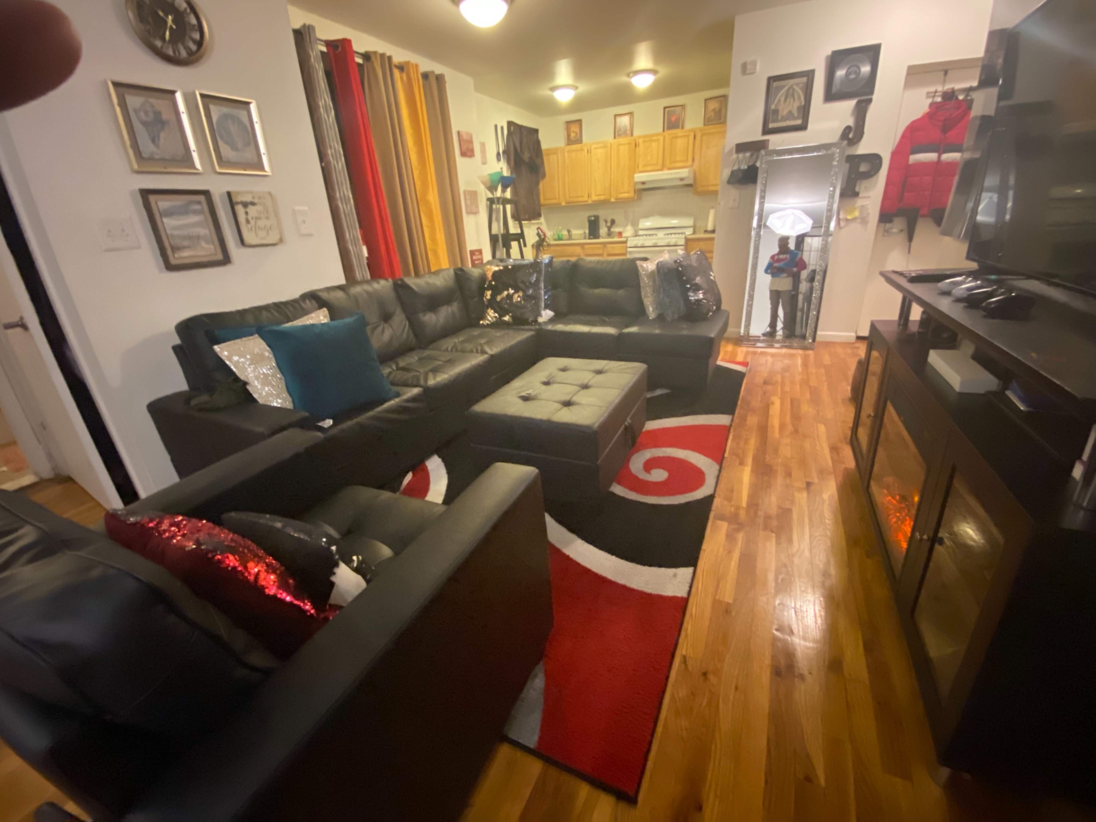 The image shows a living room with a black sectional couch, a red and black area rug, a coffee table, and a kitchen visible in the background.