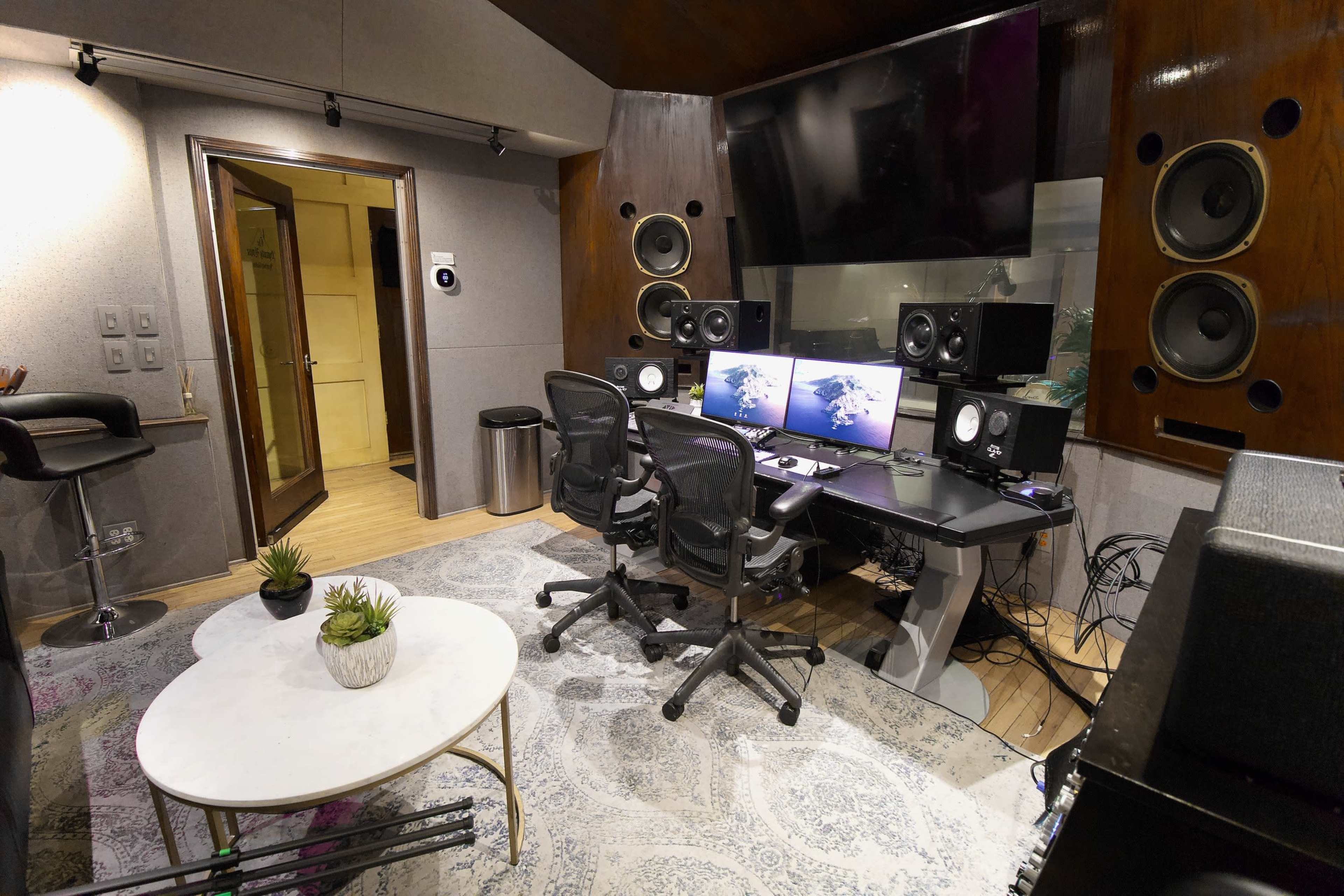 A music recording studio featuring two computer monitors, professional audio speakers, a desk with chairs, and a small round table with a plant.