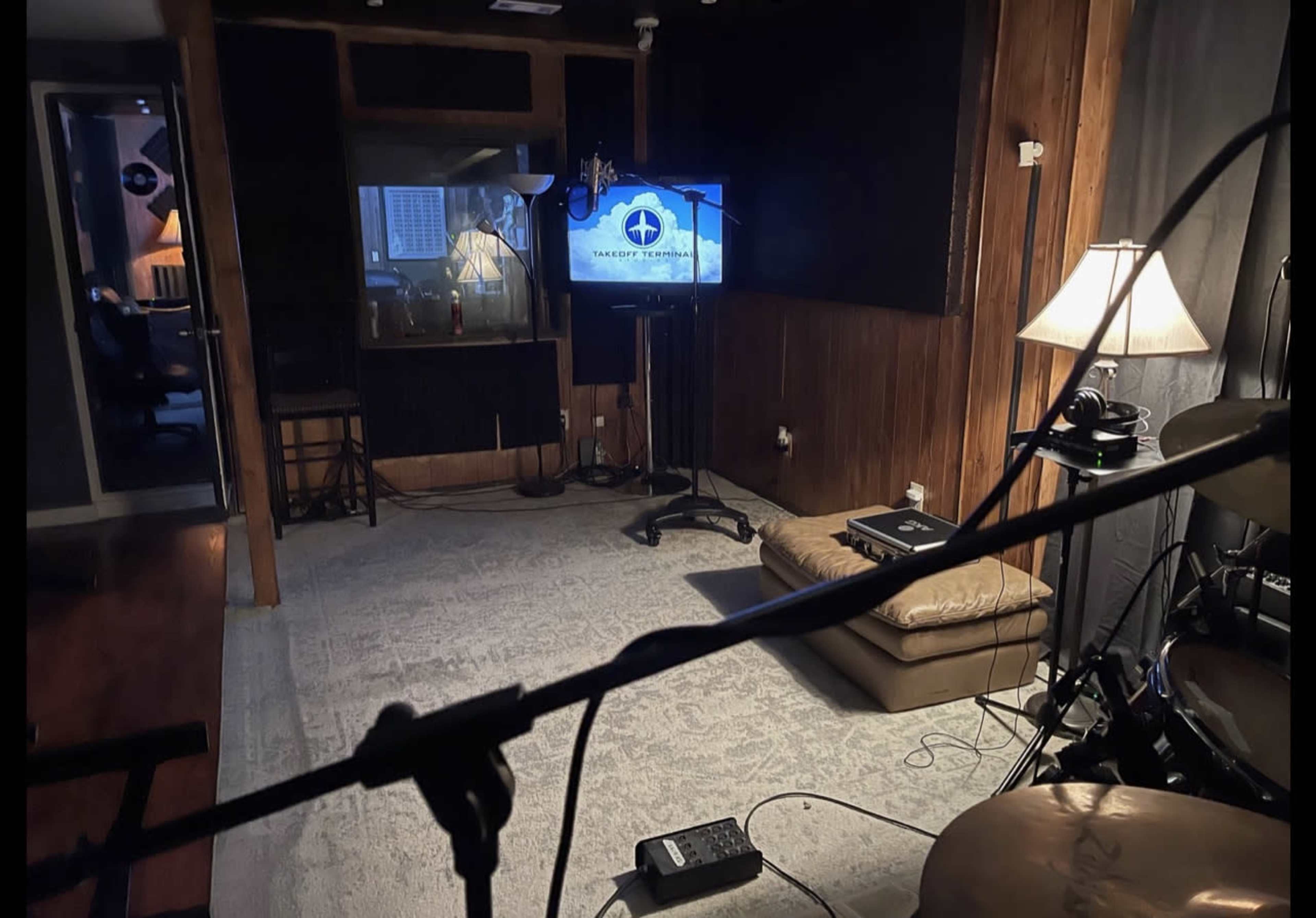A music studio features a couch, TV, and lamp, with a drum set and microphone in the foreground.