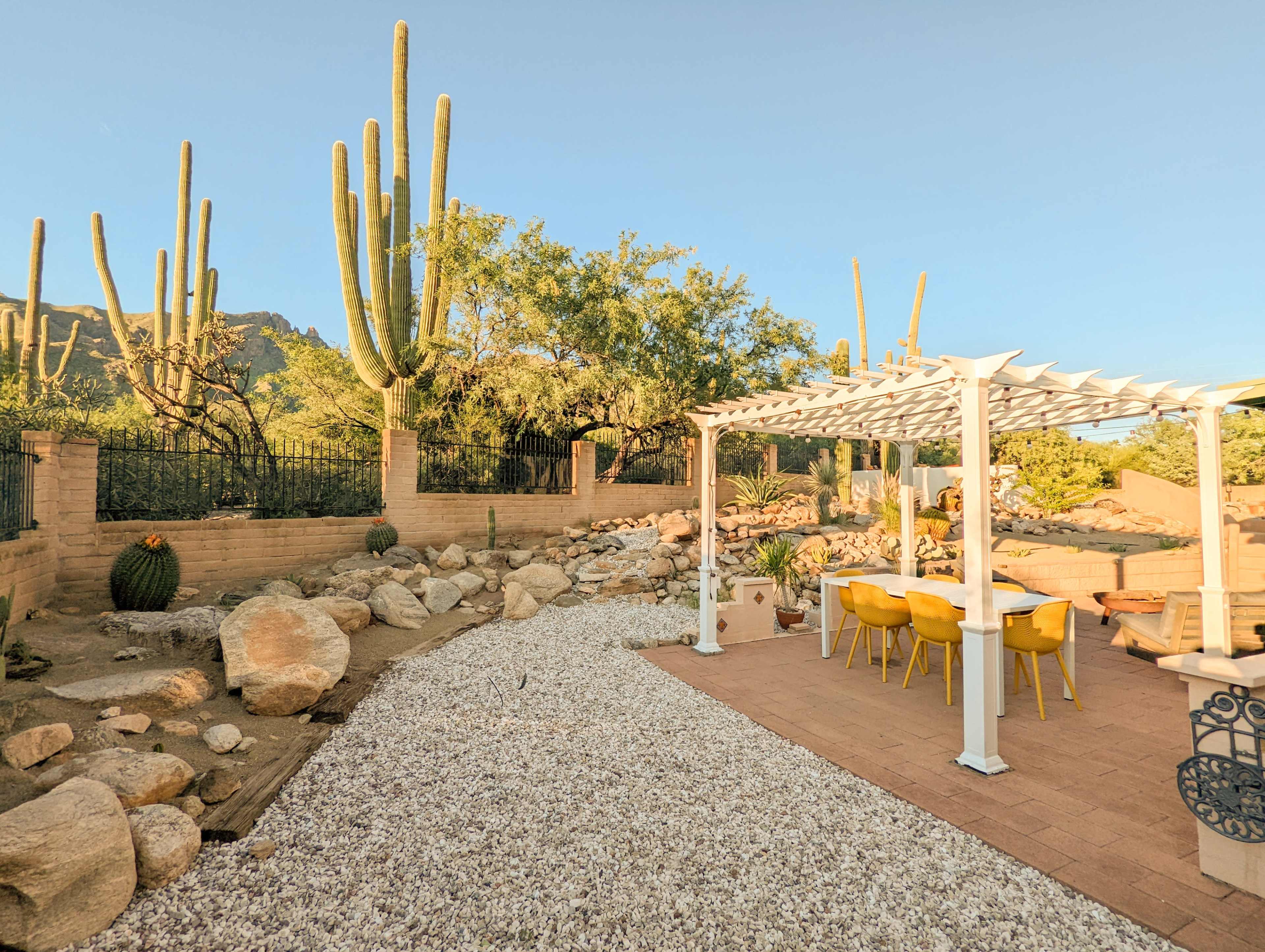 A patio area with yellow chairs under a white pergola is set against a rocky landscape featuring cacti and desert vegetation.