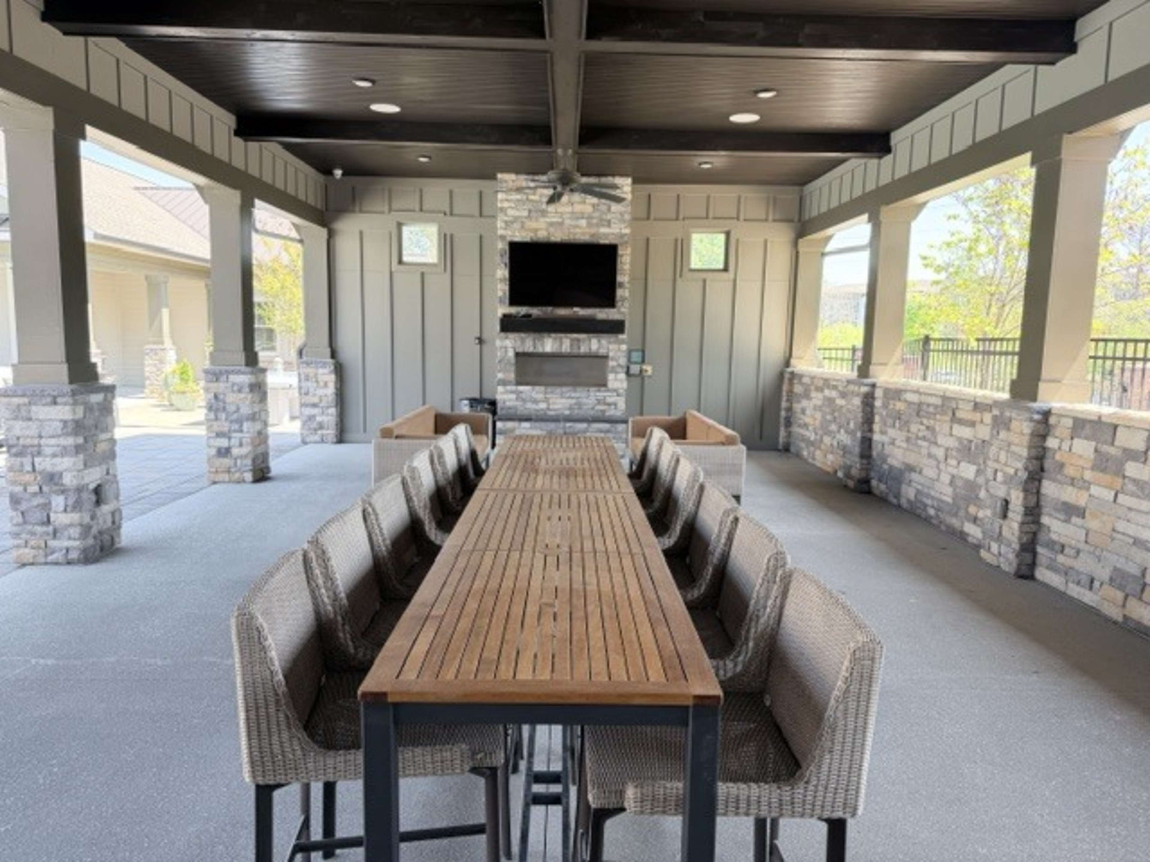 The image shows a spacious outdoor pavilion featuring a long wooden table surrounded by high-backed chairs, with stone accents and a central fireplace area.