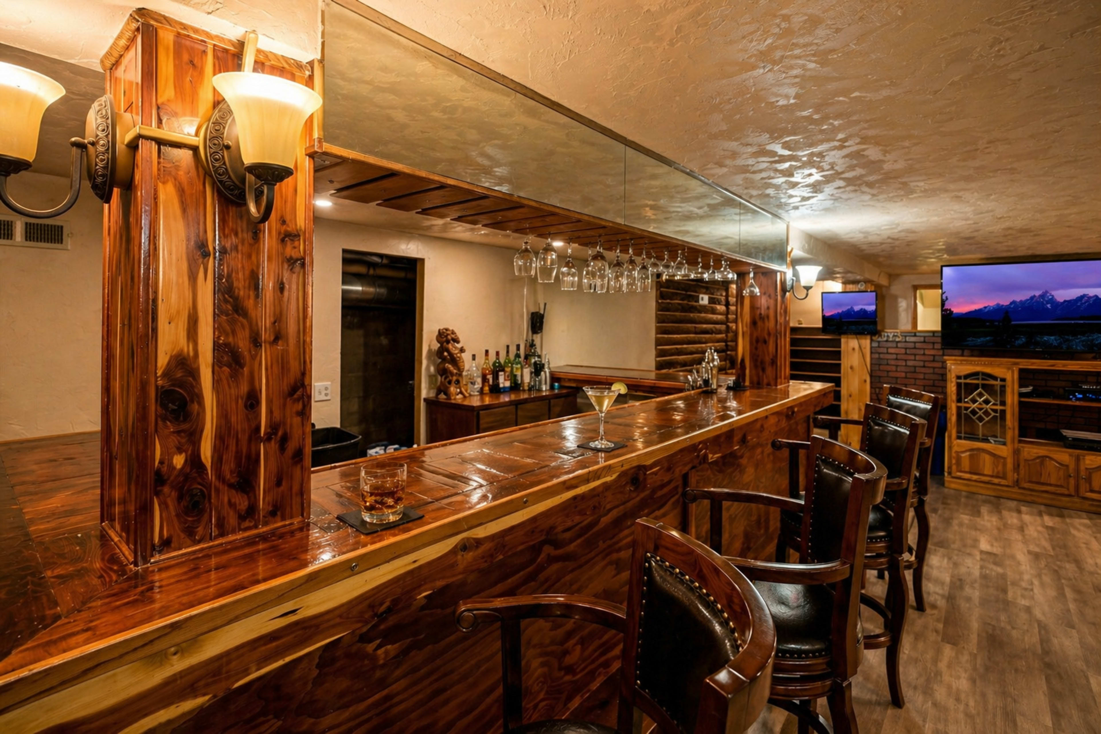The image shows a wooden bar area with high stools, bottles on the shelves, and a television mounted on the wall.