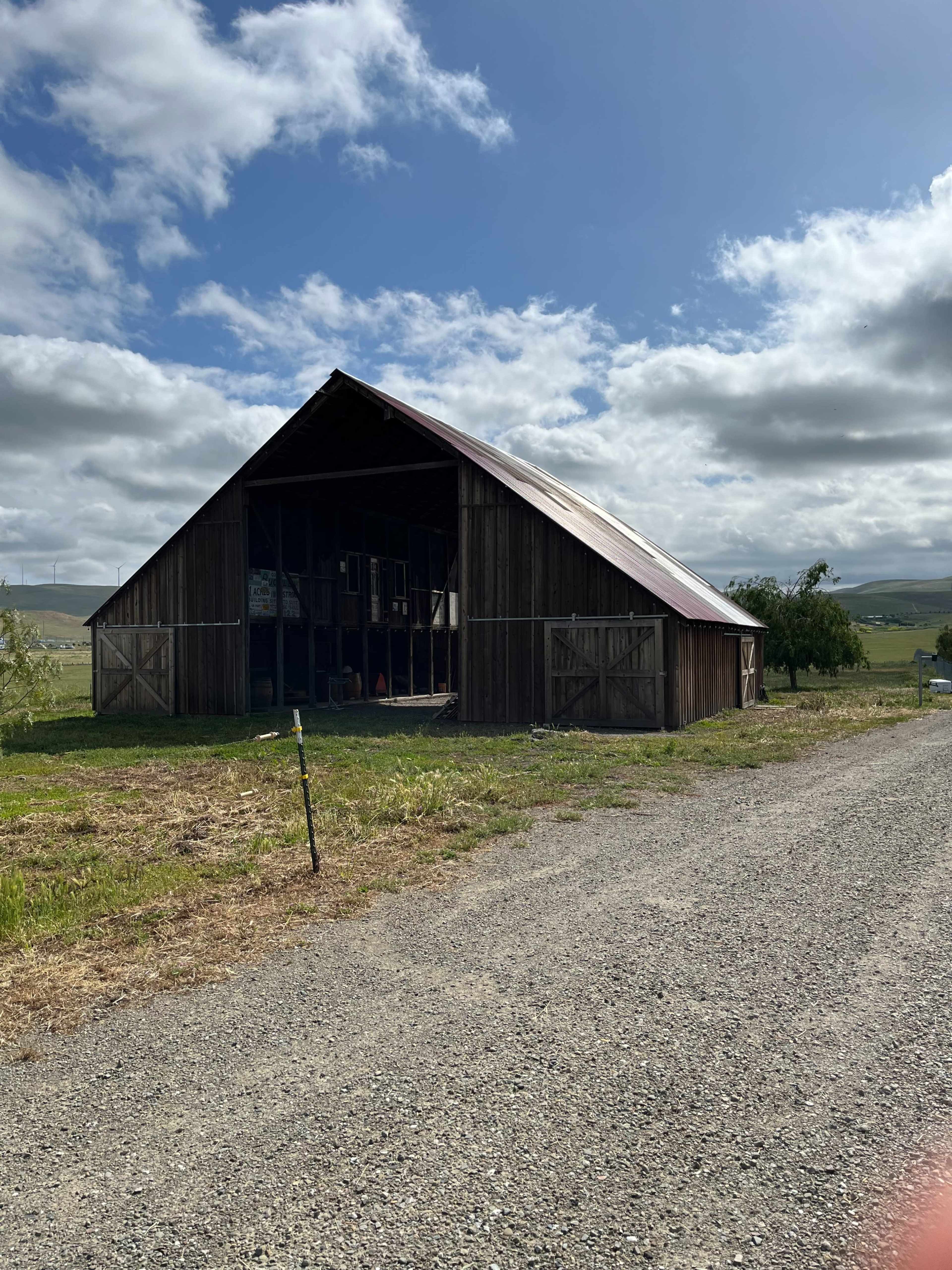 A wooden barn with a metal roof is situated next to a gravel road under a cloudy sky.