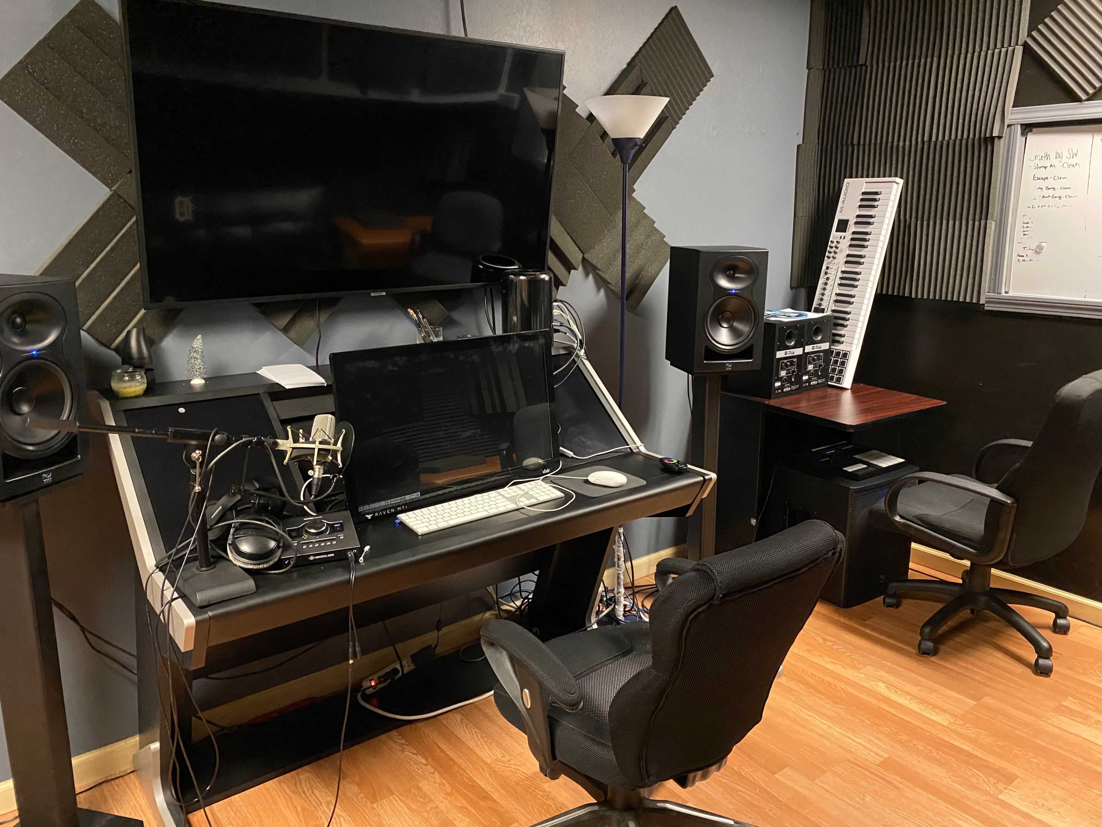 A music production studio featuring a desk with a computer, speakers, and a microphone, along with a keyboard and additional equipment on the walls.
