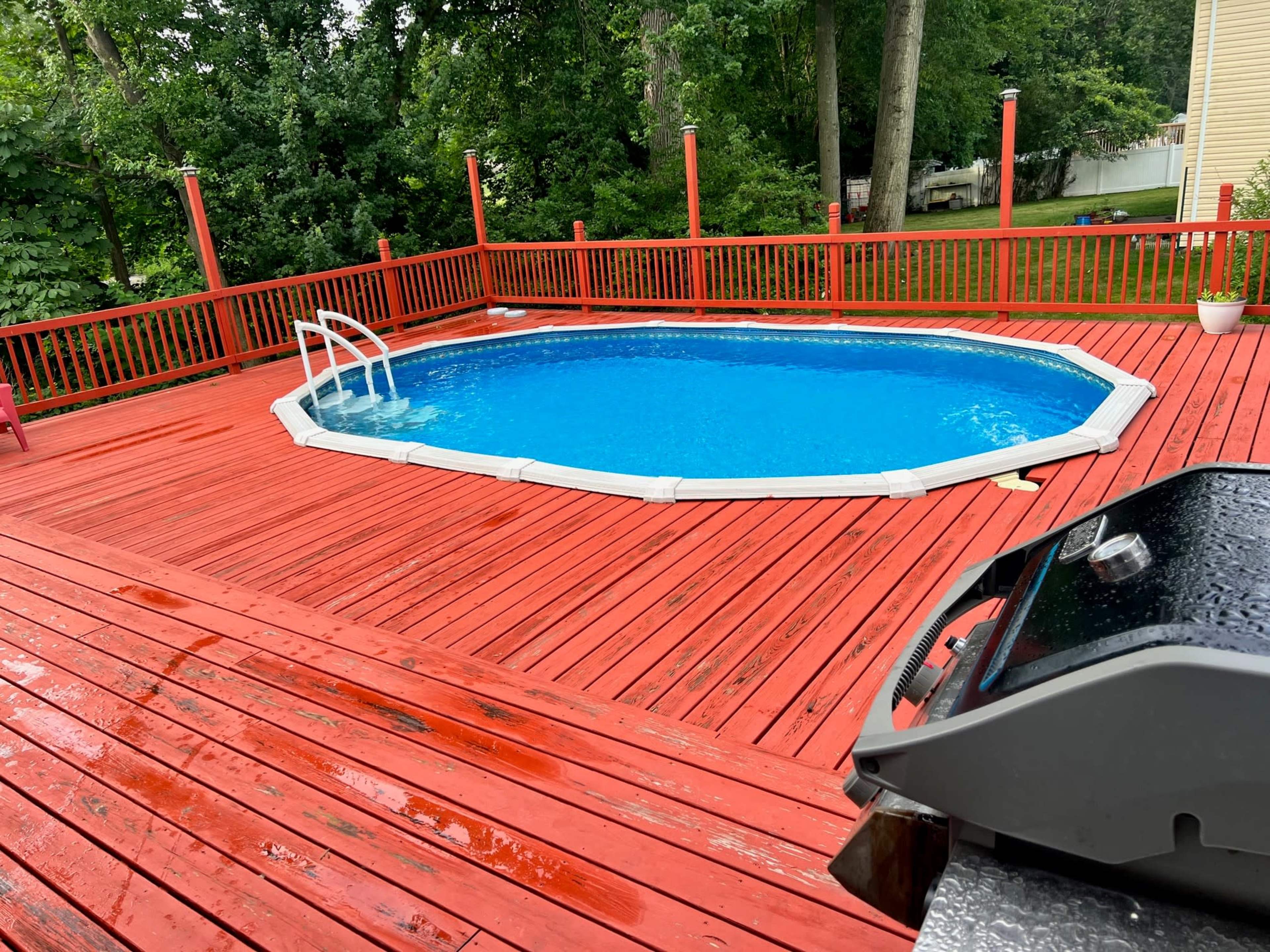 A red wooden deck surrounds an oval swimming pool in a green backyard.