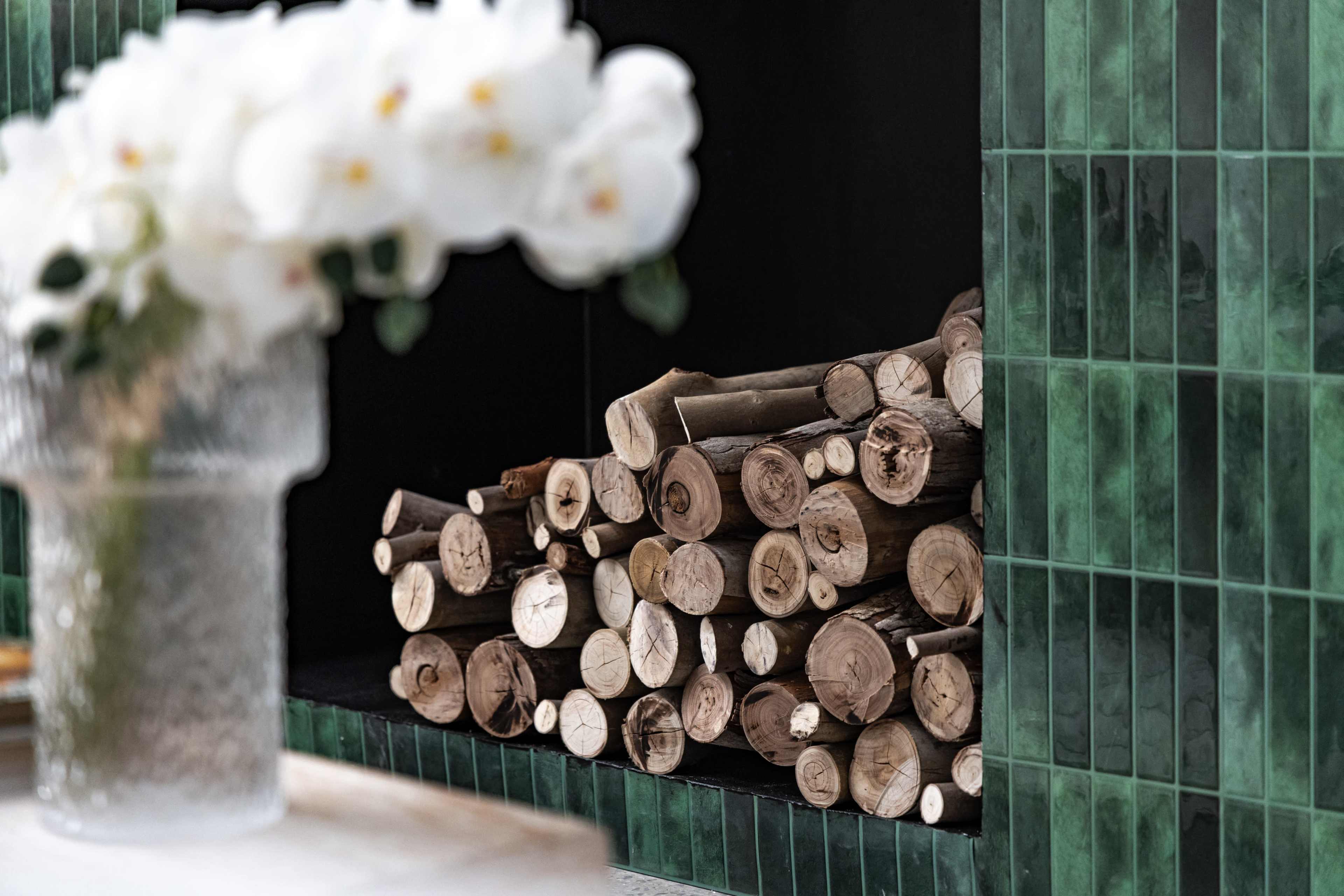 The image shows a neatly stacked pile of wooden logs beside a vase of white flowers set against a green tiled wall.