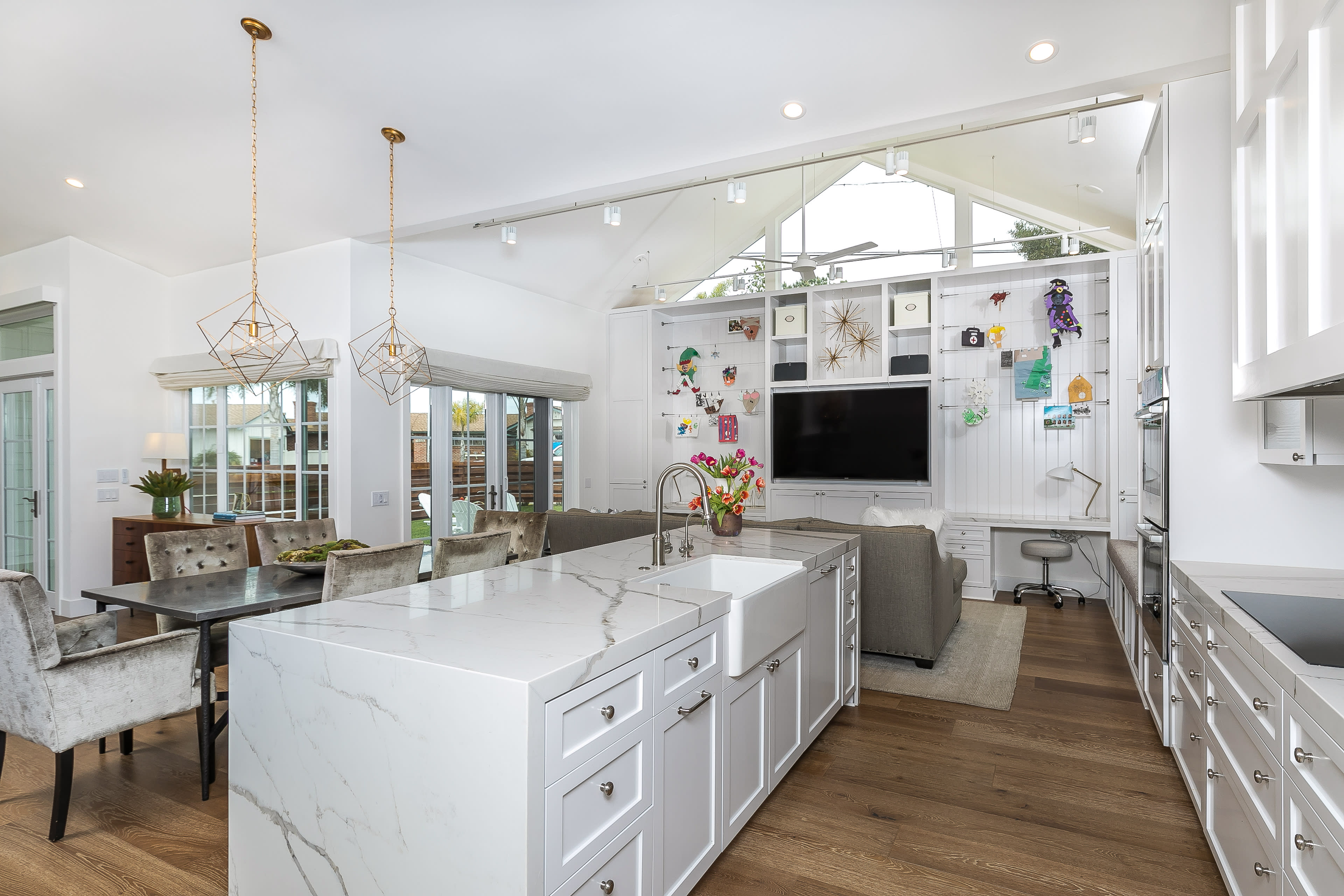 A modern kitchen features a large island with white countertops, surrounded by a dining area and a living space with large windows and a wall-mounted television.