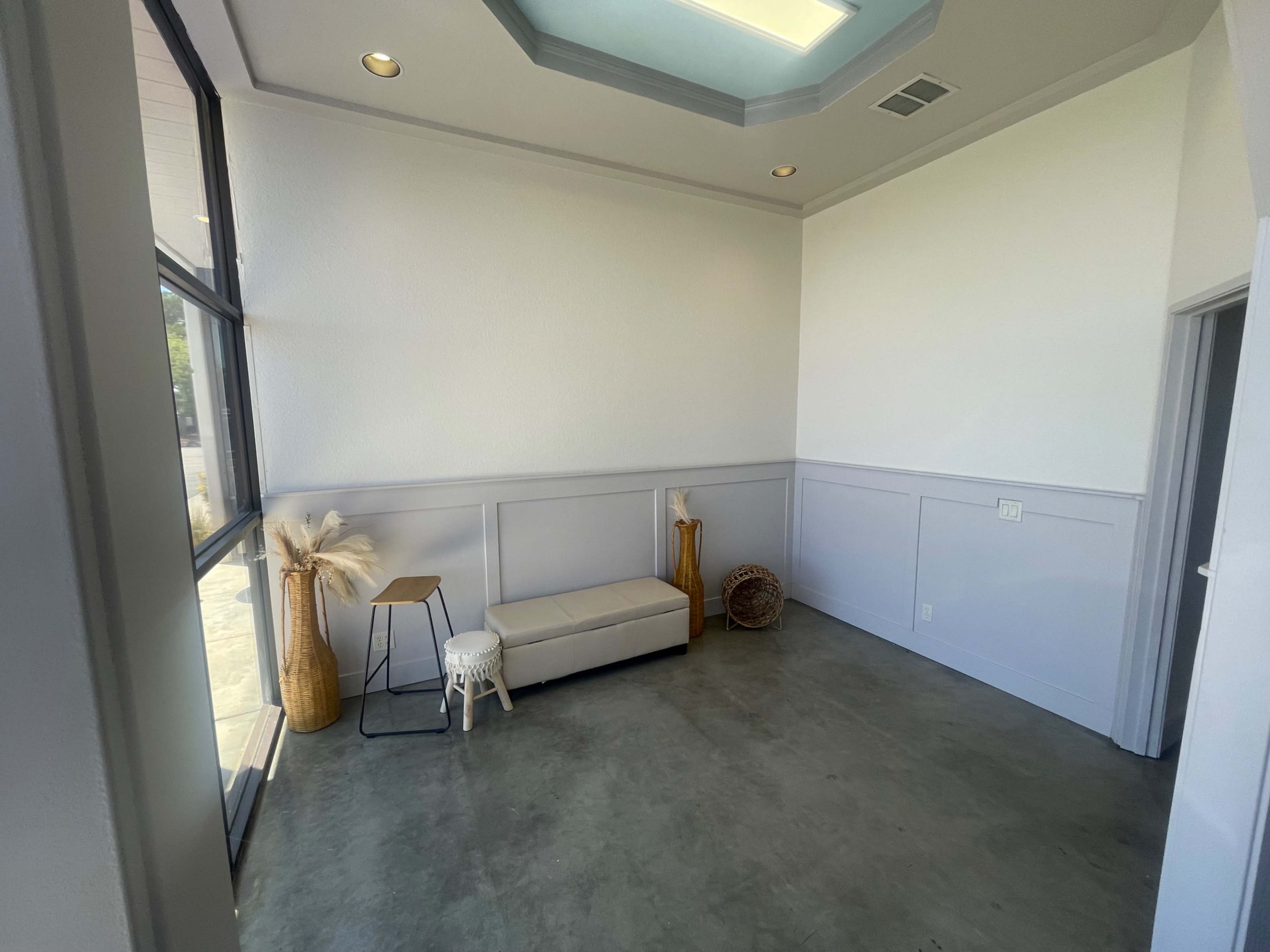 The image shows a sparsely furnished interior room with a bench, a small stool, and decorative items, featuring large windows that let in natural light.