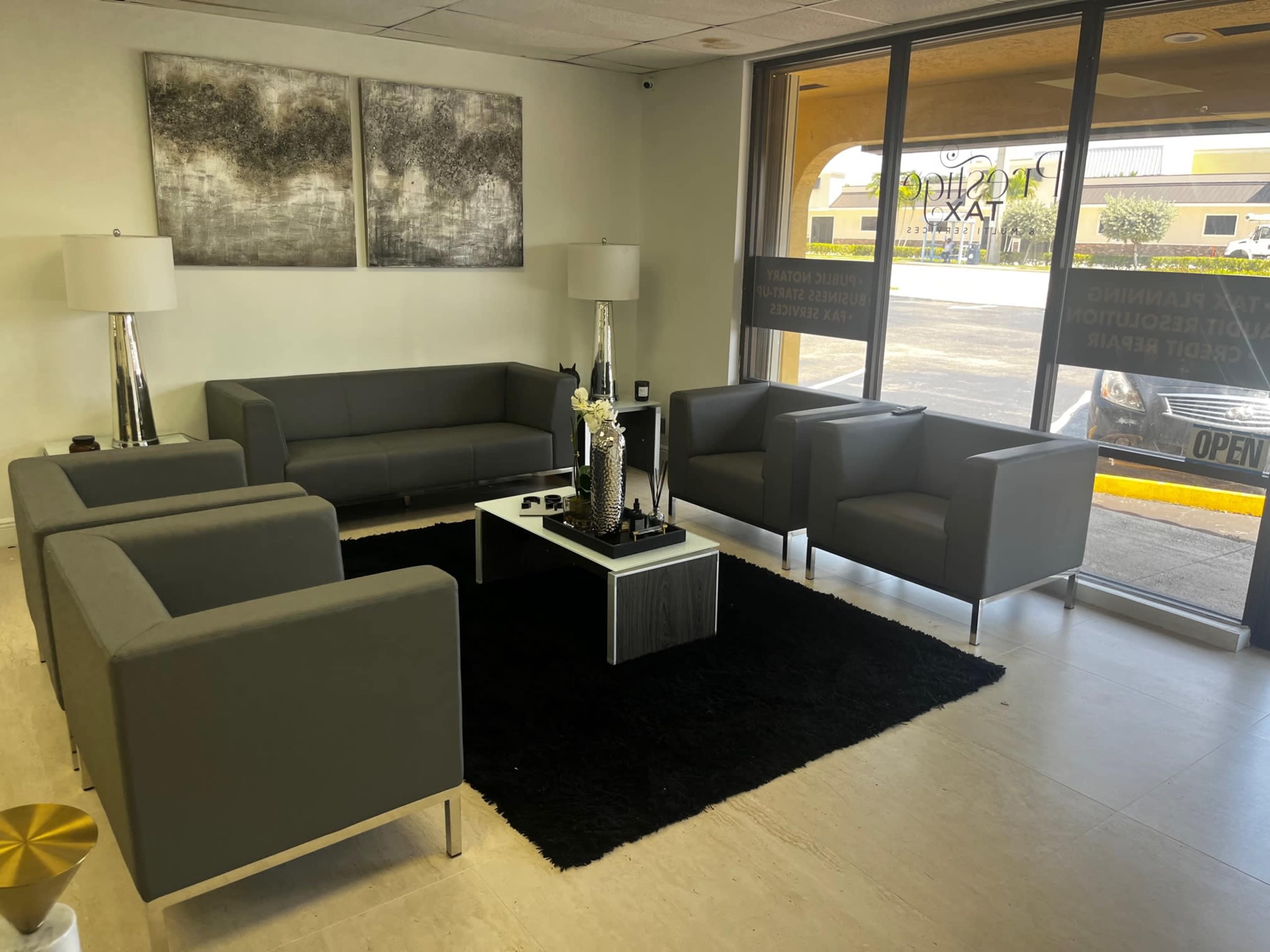 A modern waiting area with gray sofas and chairs arranged around a coffee table on a black rug, with large windows letting in natural light.