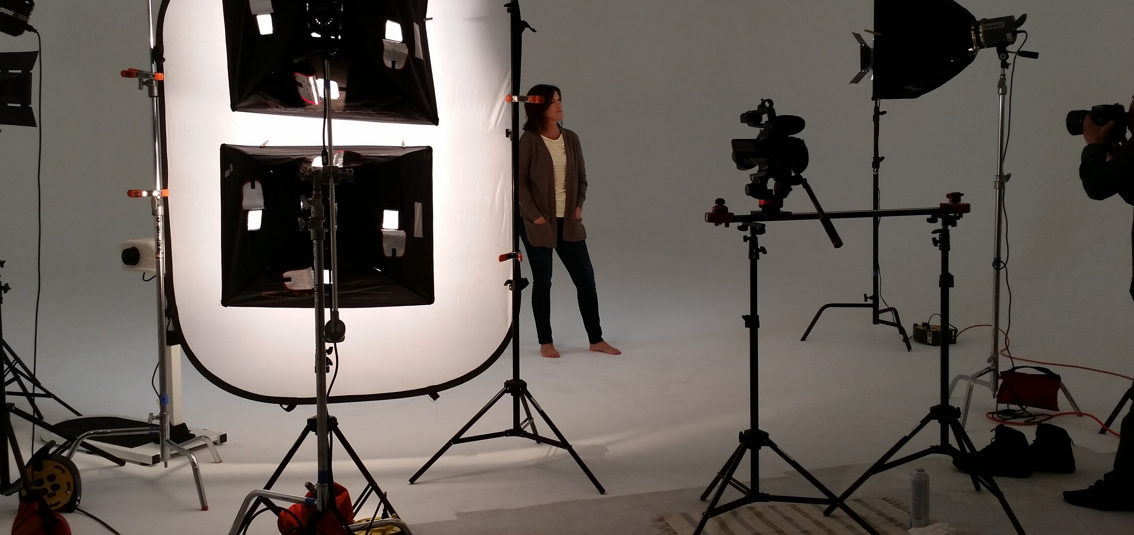 A model stands barefoot in a well-lit photography studio surrounded by multiple lights and cameras.