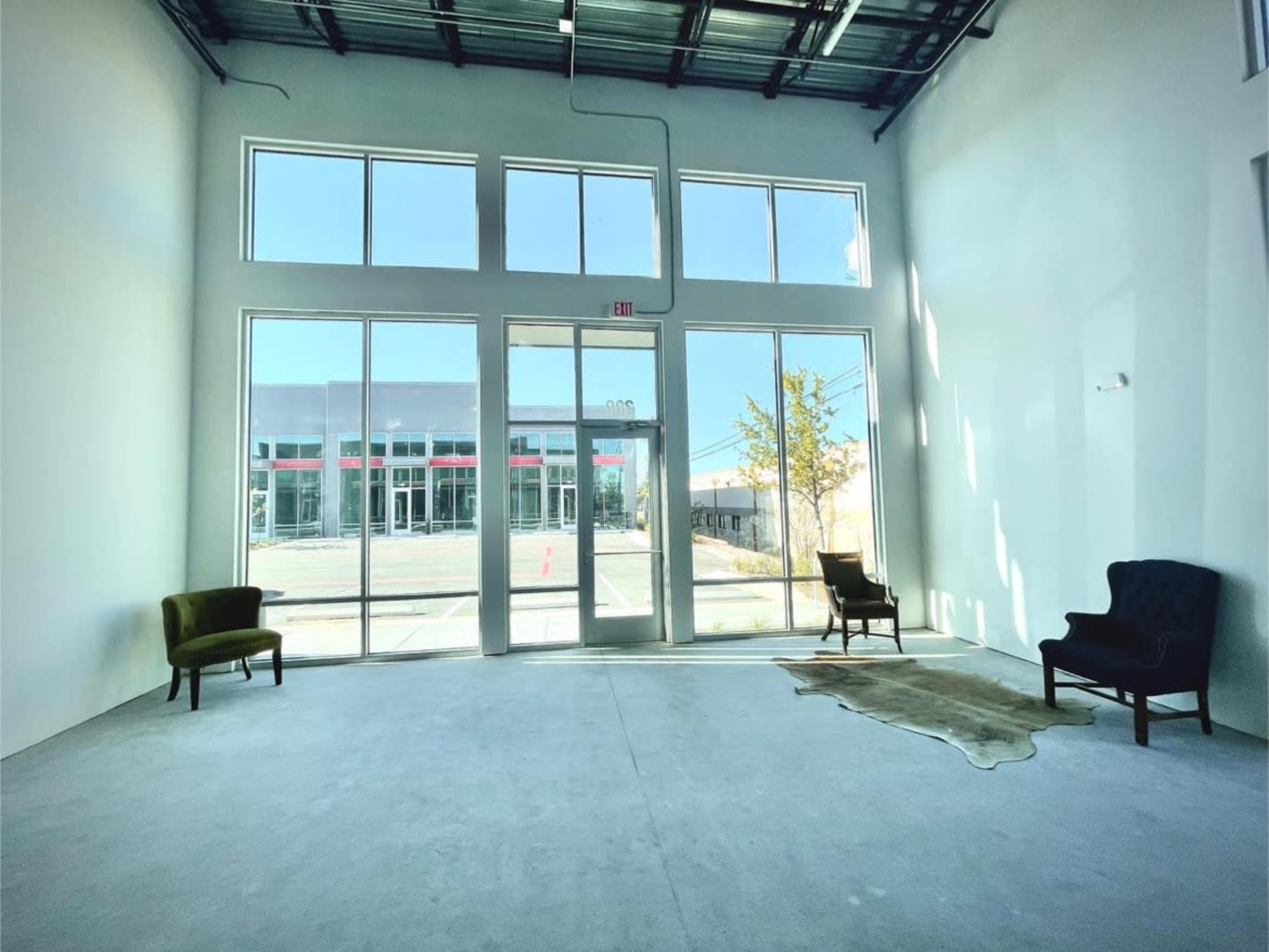 A spacious, empty interior with large windows and two chairs flanking a rug on a concrete floor.