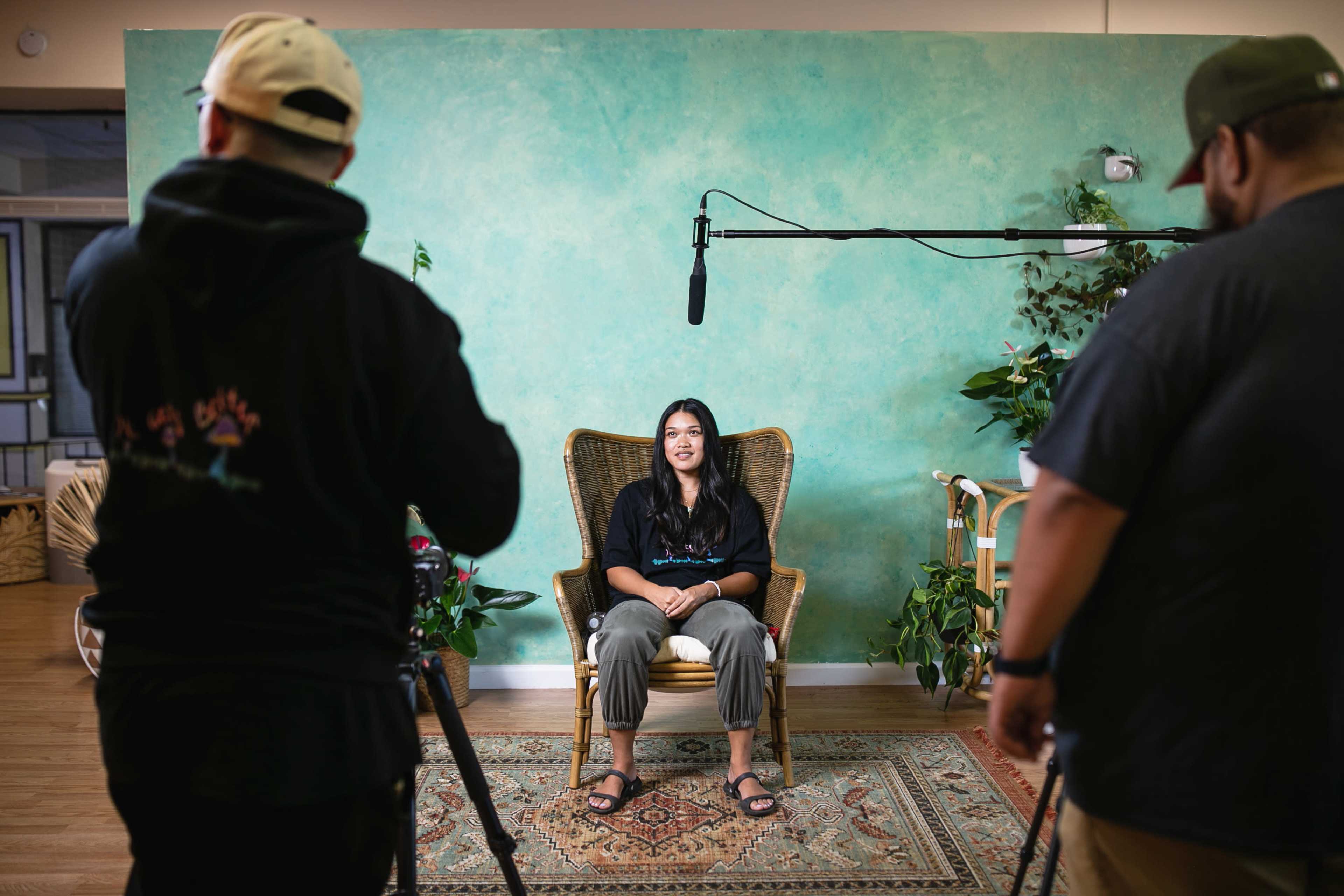A woman sits in a wicker chair in front of a green wall while two people prepare for filming with cameras and a microphone.