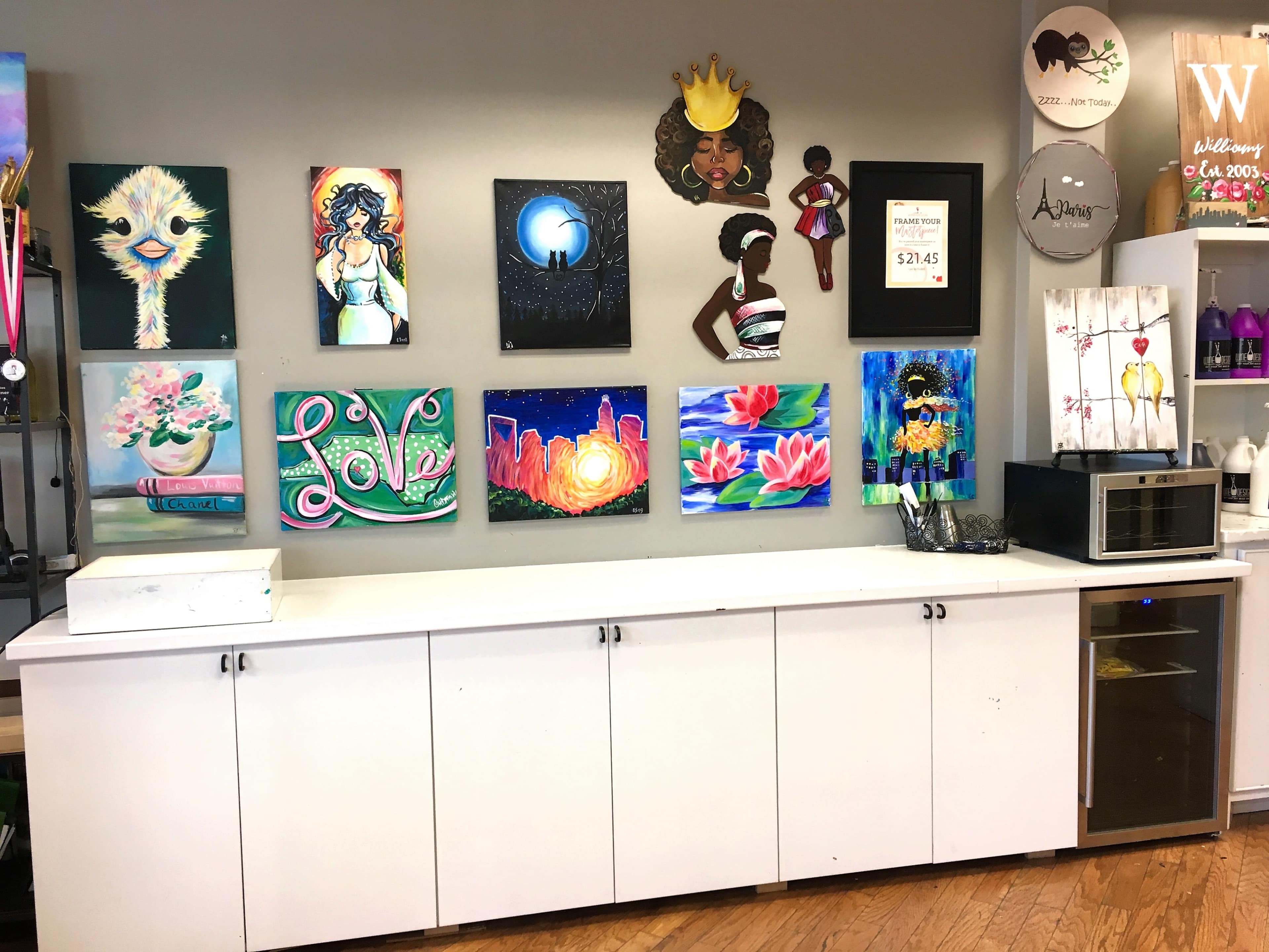 A wall adorned with various colorful paintings, including portraits and abstract designs, above a white cabinet with a microwave and a drink cooler.