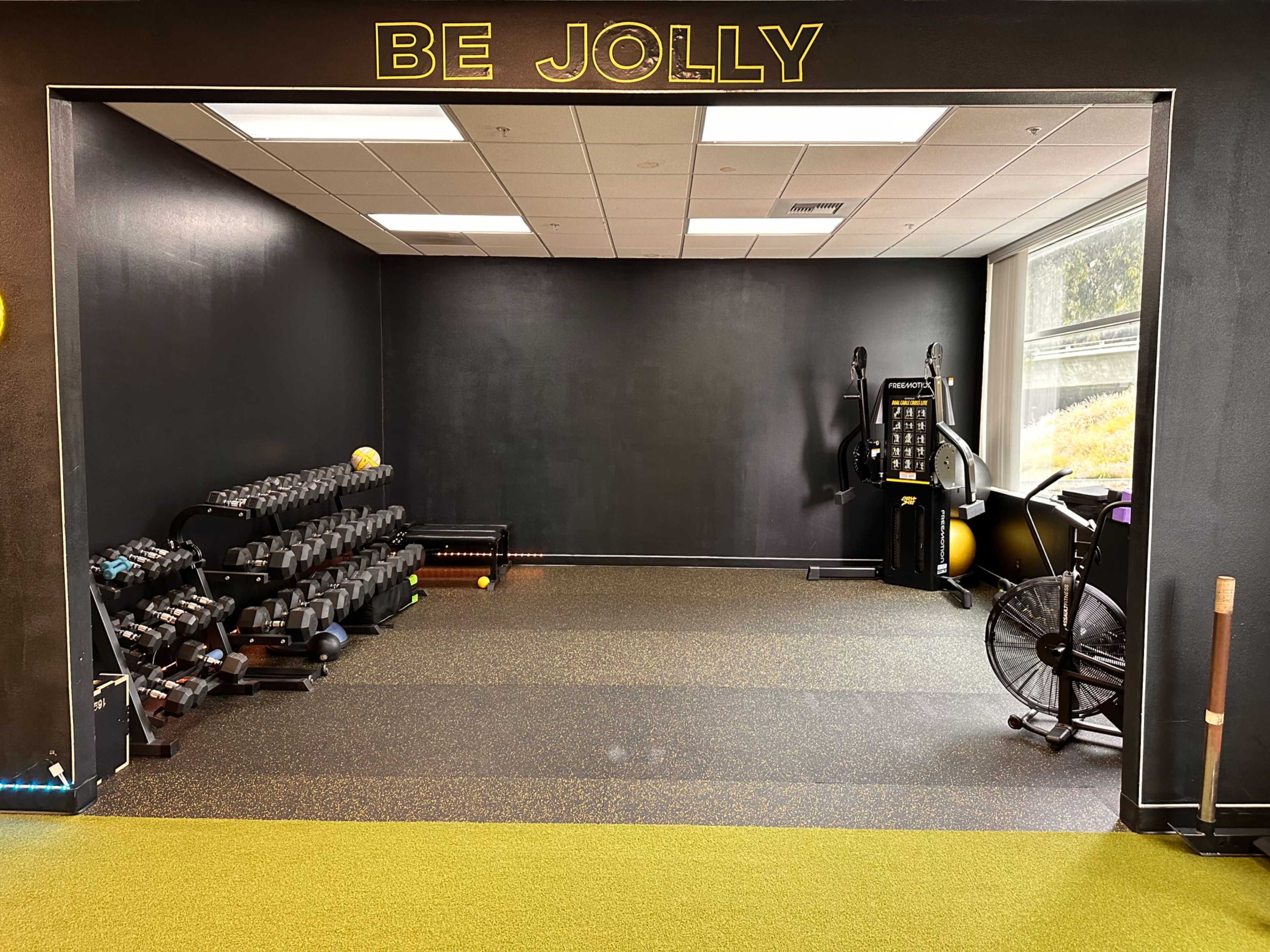A fitness area with weights on racks, a stationary bike, and exercise equipment against a dark wall featuring the words "BE JOLLY."