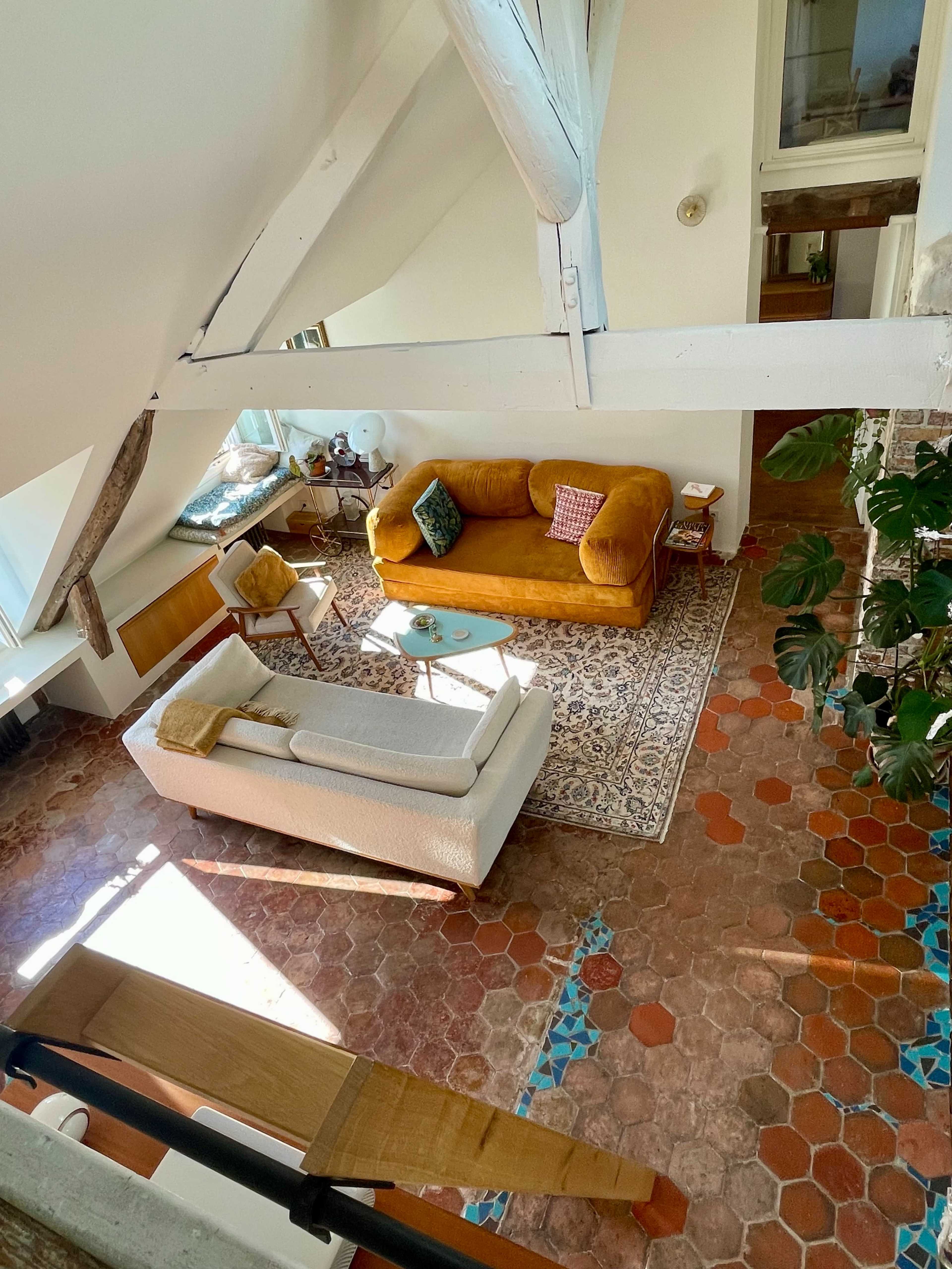 The image shows a spacious living room with a yellow couch, a white sofa, a decorative rug, and a variety of plants, set on terracotta tiled flooring.