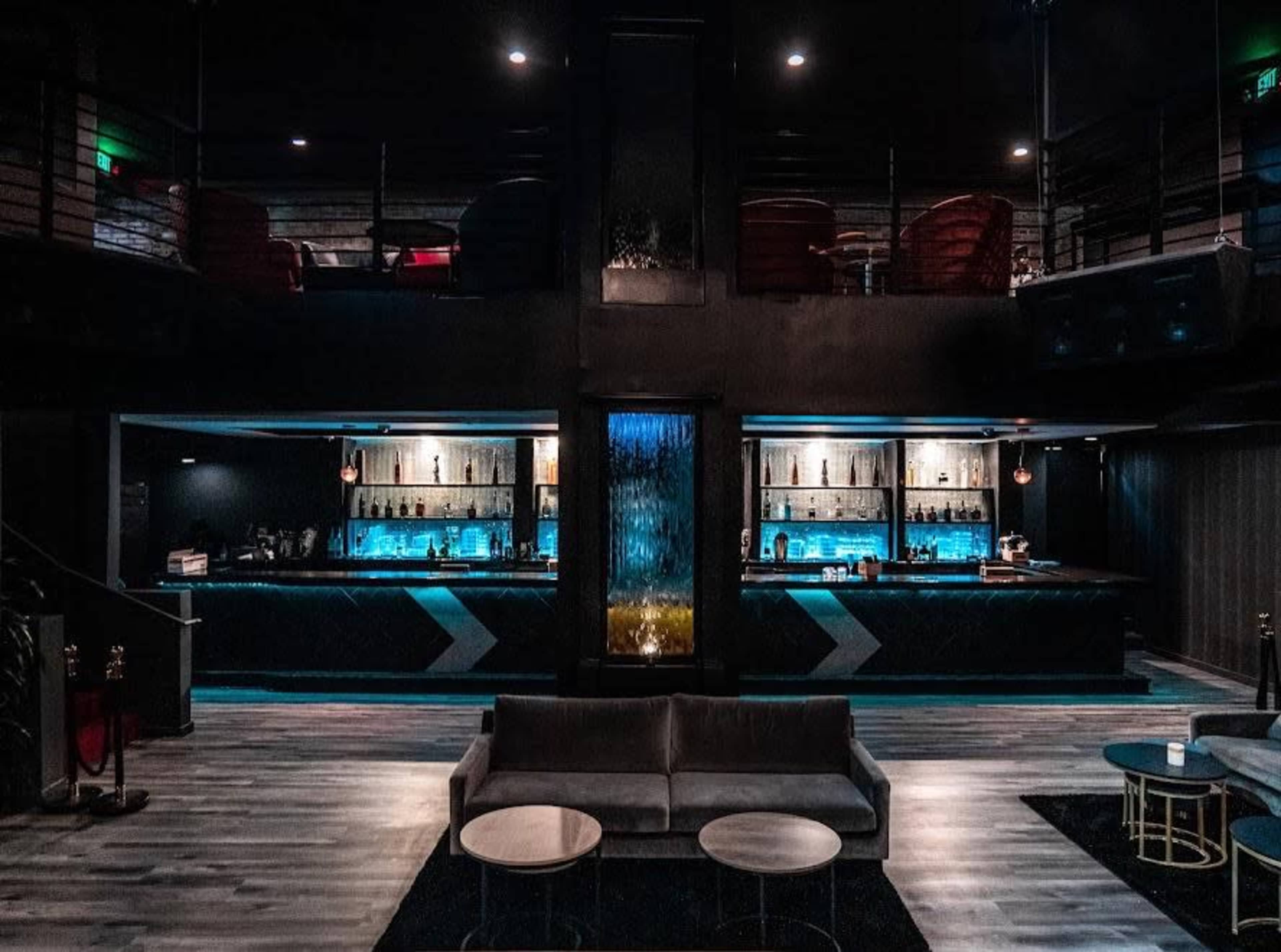 A dimly lit bar area with a waterfall feature, a long illuminated bar, and seating arranged on two levels.