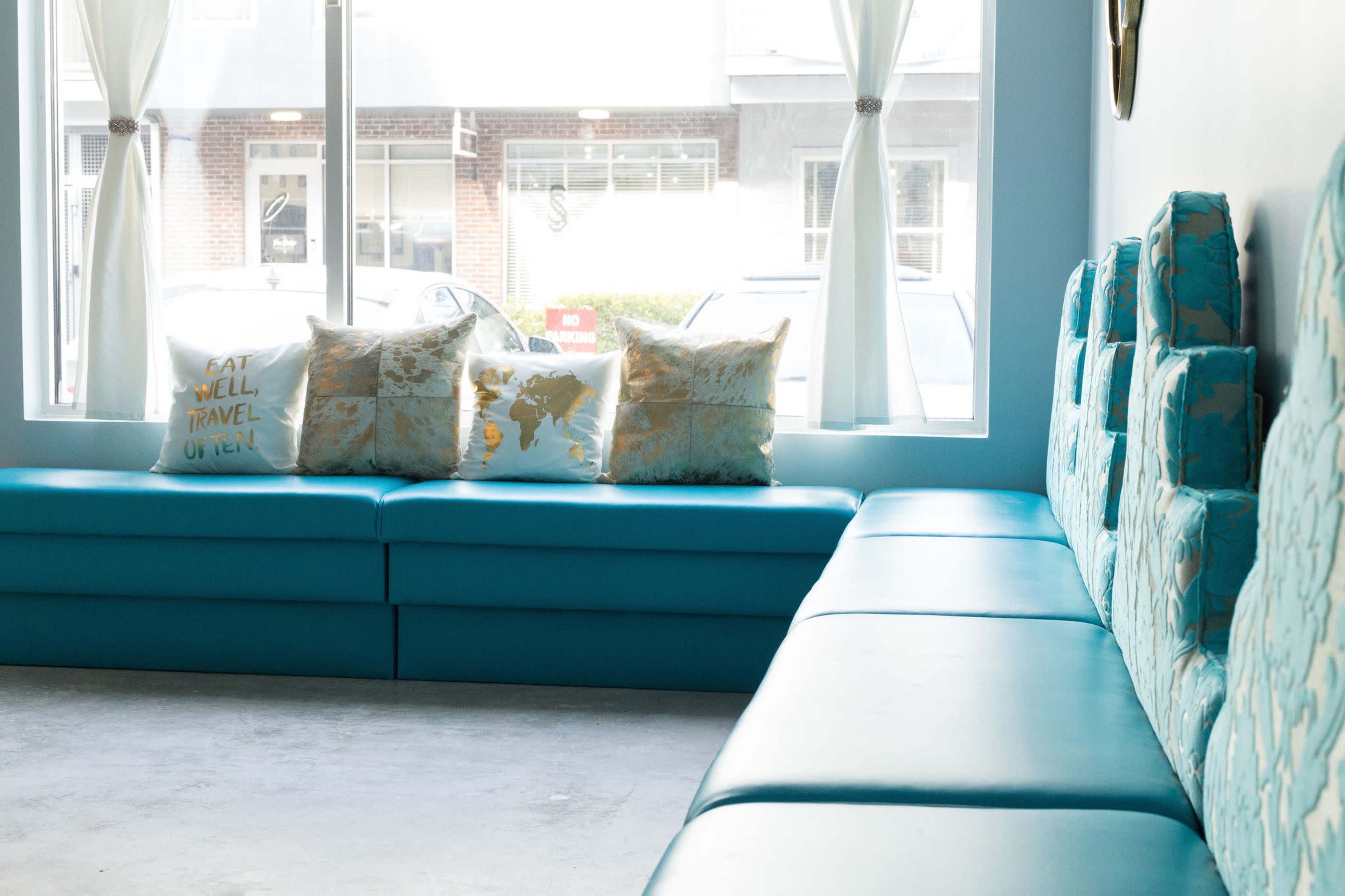 The image shows a cozy seating area with a light blue bench, patterned cushions, and large windows letting in natural light.
