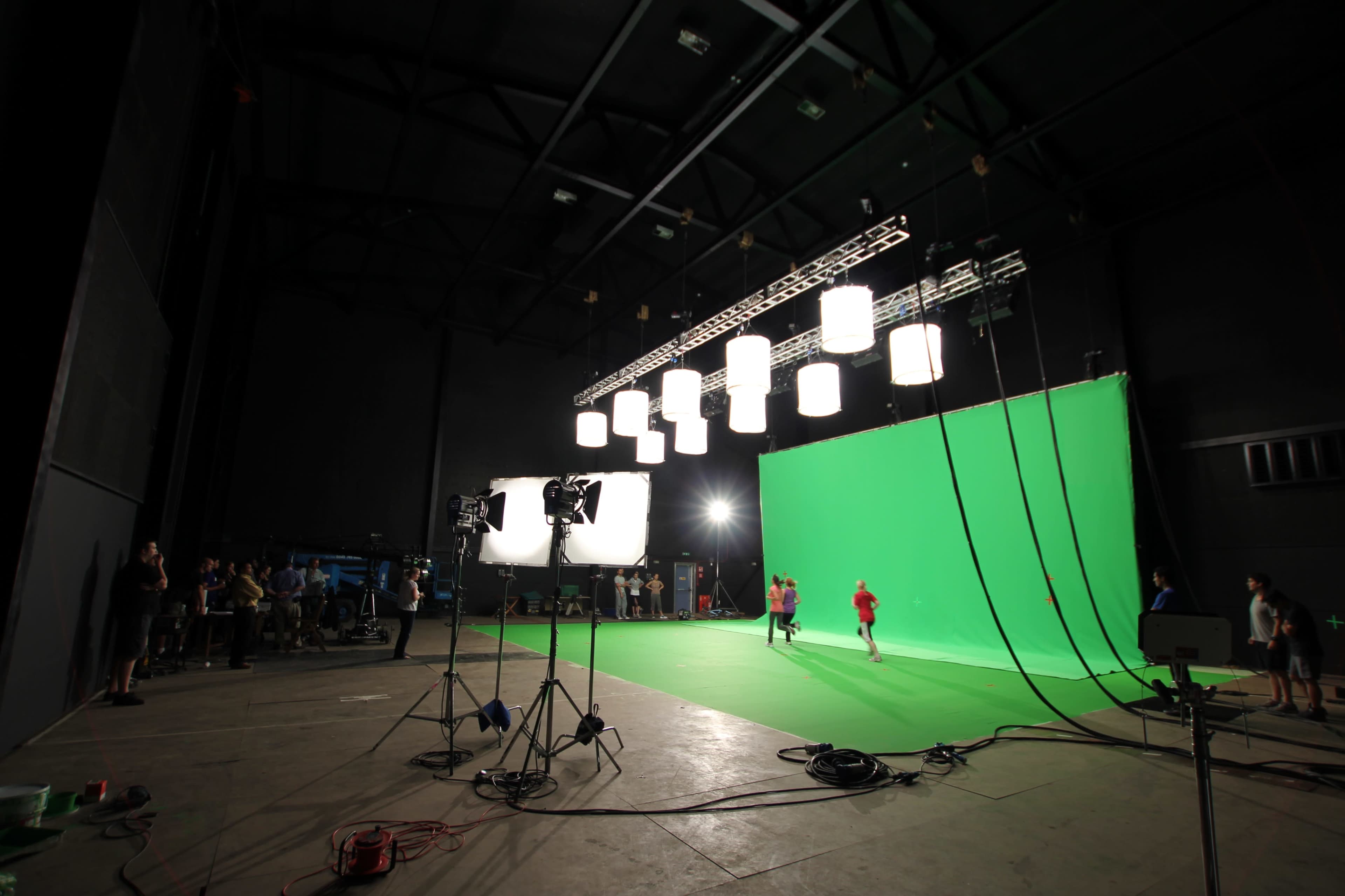 A large studio with a green screen backdrop, bright lights, and individuals performing in the foreground.