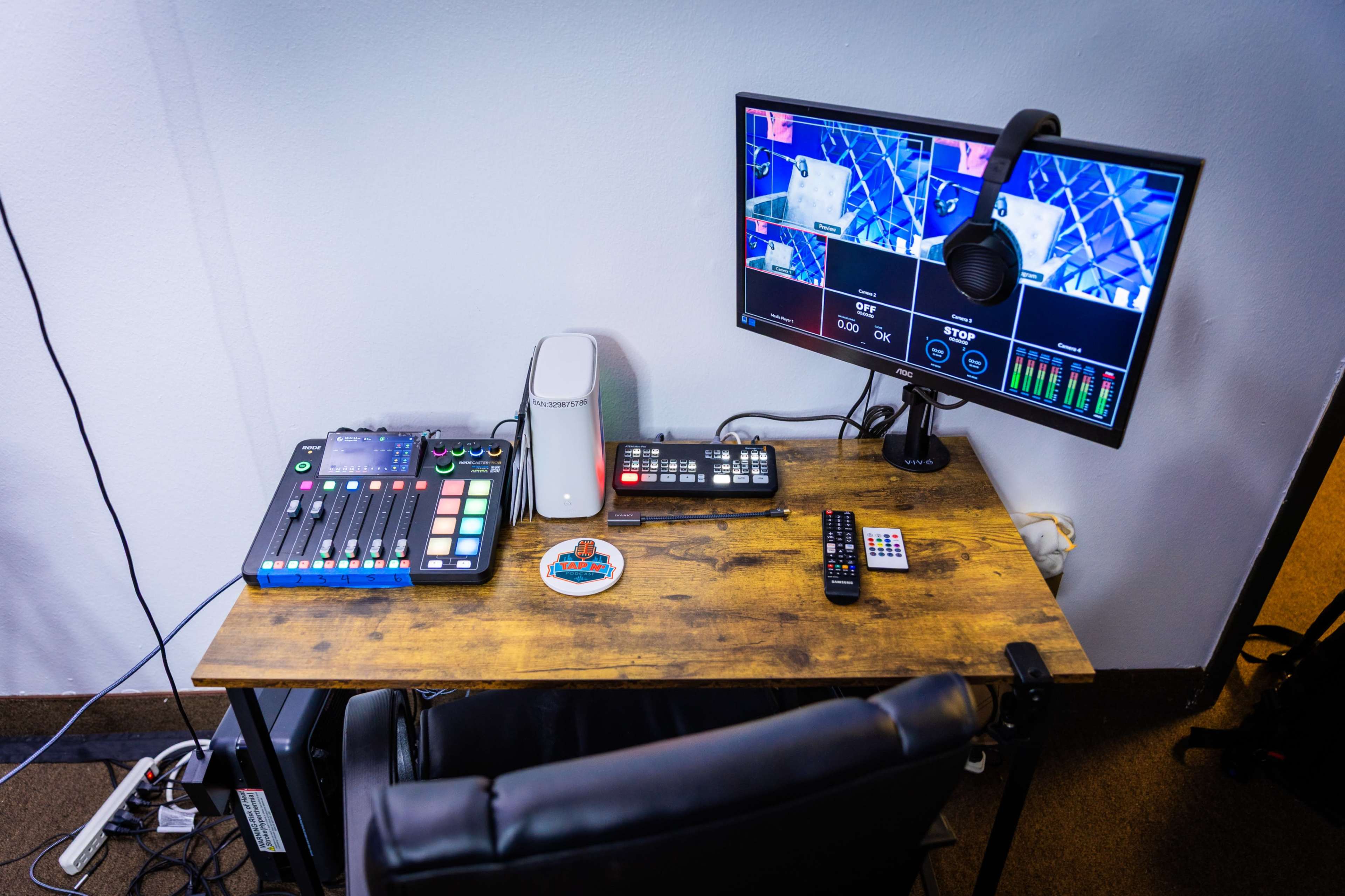 The image shows a modern broadcasting setup with a control panel, a computer monitor displaying video feeds, and a swivel chair on a wooden desk.