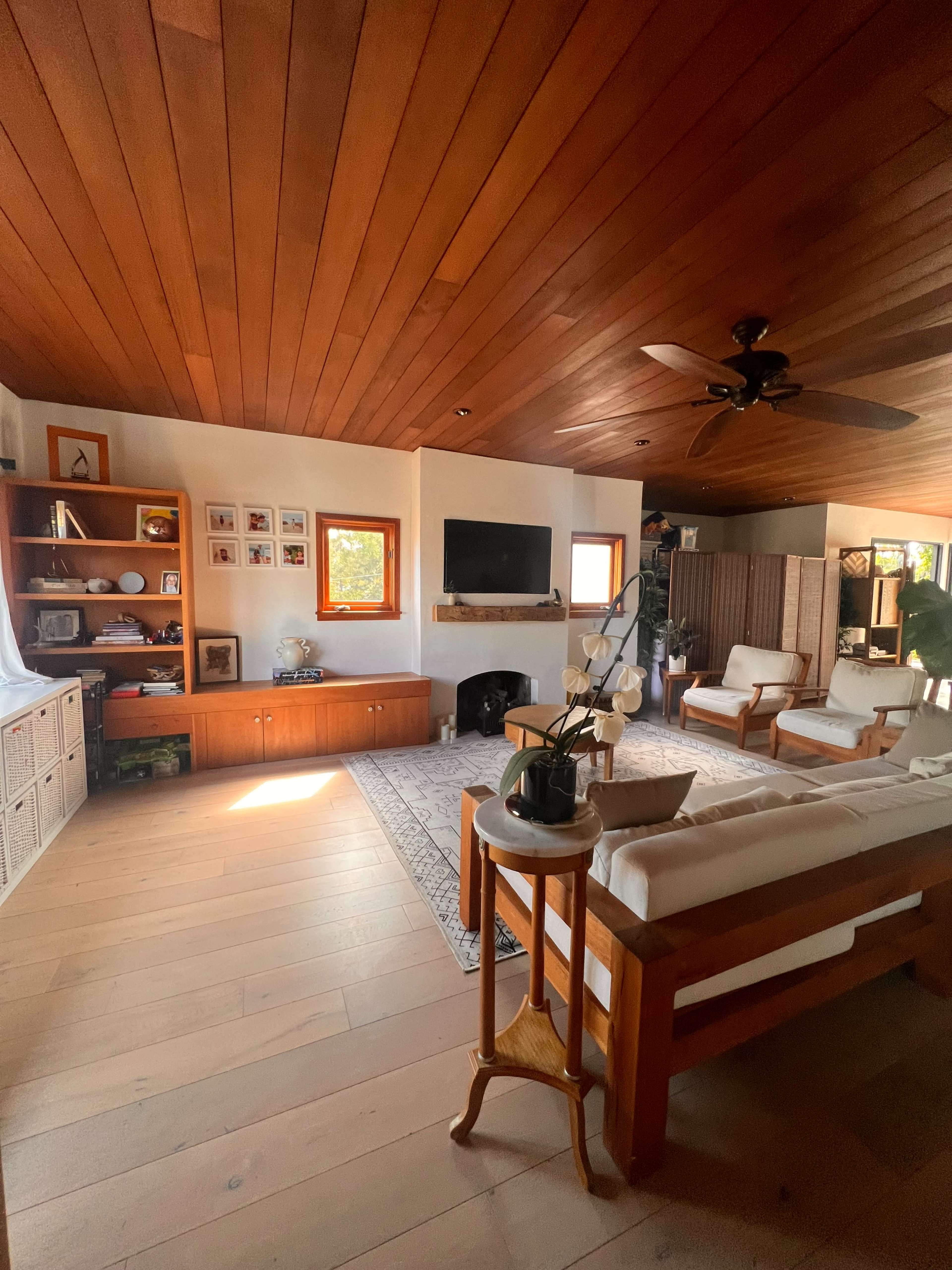 A bright living room featuring wooden walls and ceiling, a seating area with two chairs and a couch, a television mounted on the wall, and a decorative bookshelf.
