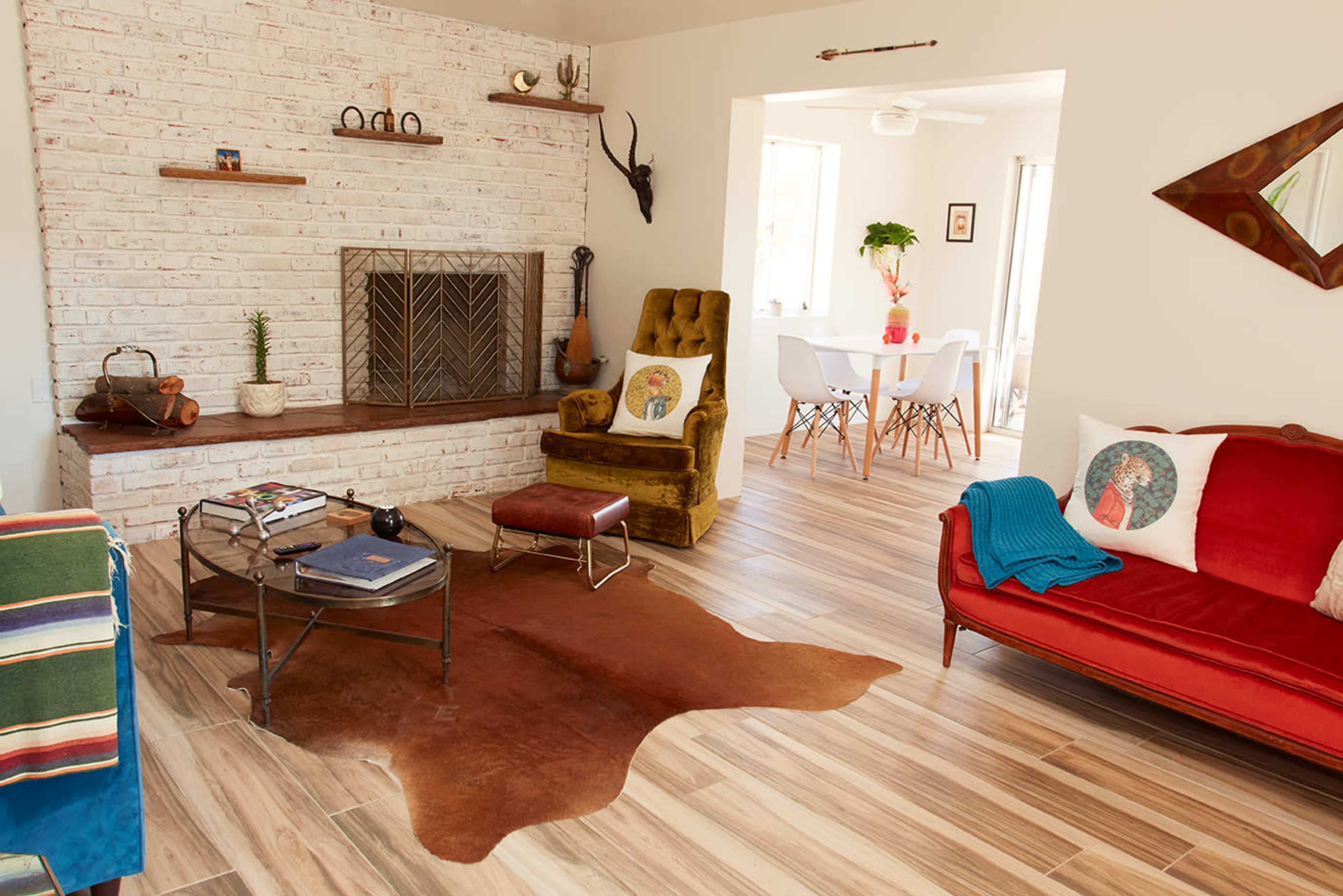 The image shows a modern living room featuring a brick fireplace, a wooden floor, a red sofa, a yellow armchair, and a dining area in the background.