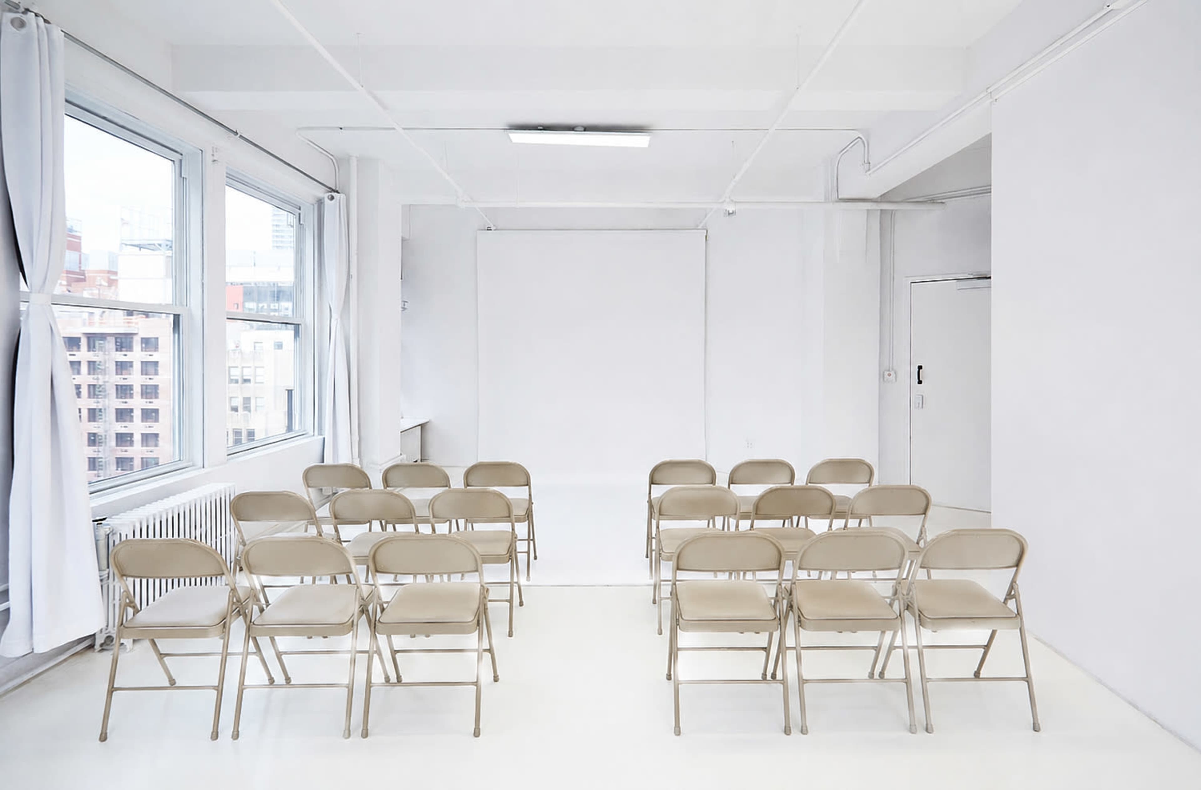 The image shows a bright, empty room with white walls and a row of beige folding chairs arranged for an event.