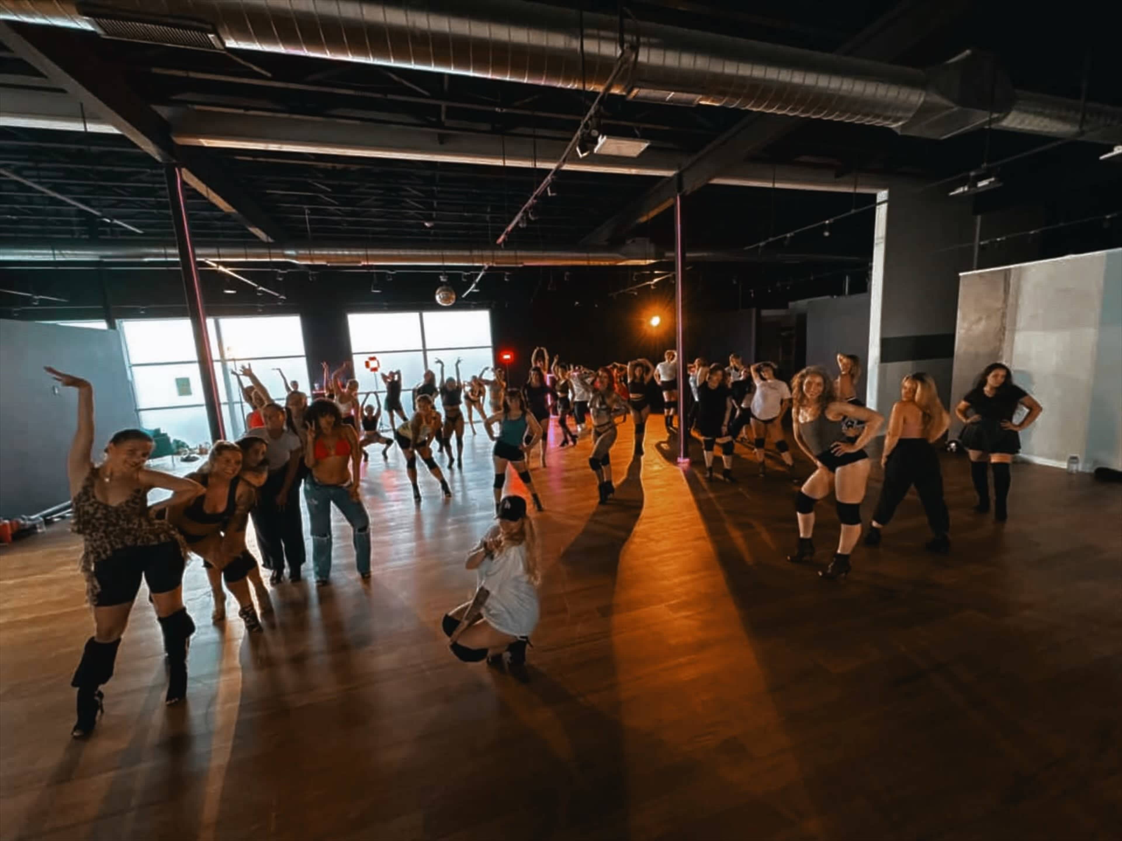 A large group of dancers is practicing in a bright studio with mirrored walls and wooden floors.