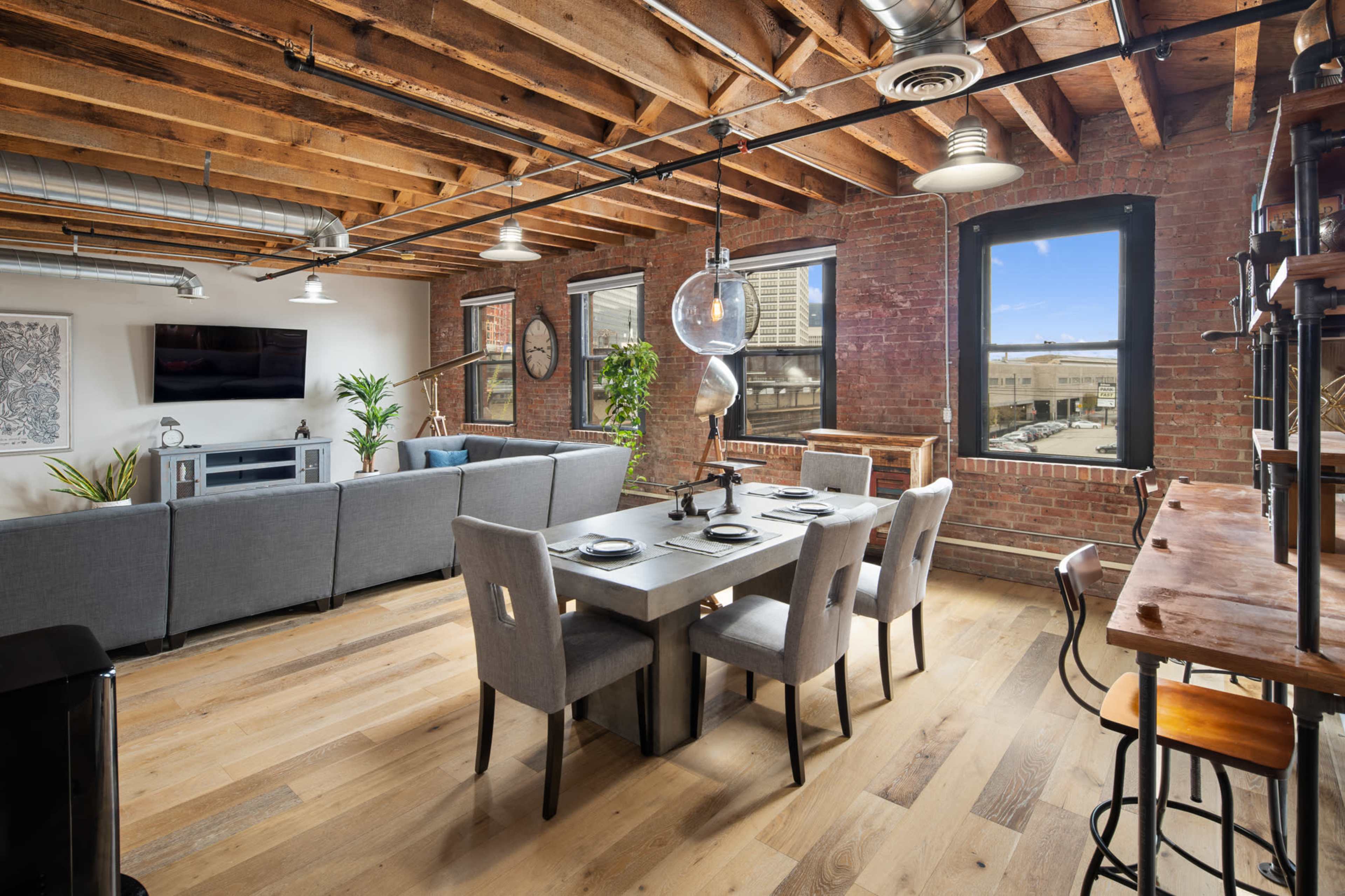 A modern open-concept living space featuring a dining table, seating area with a television, and large windows allowing natural light to enter.