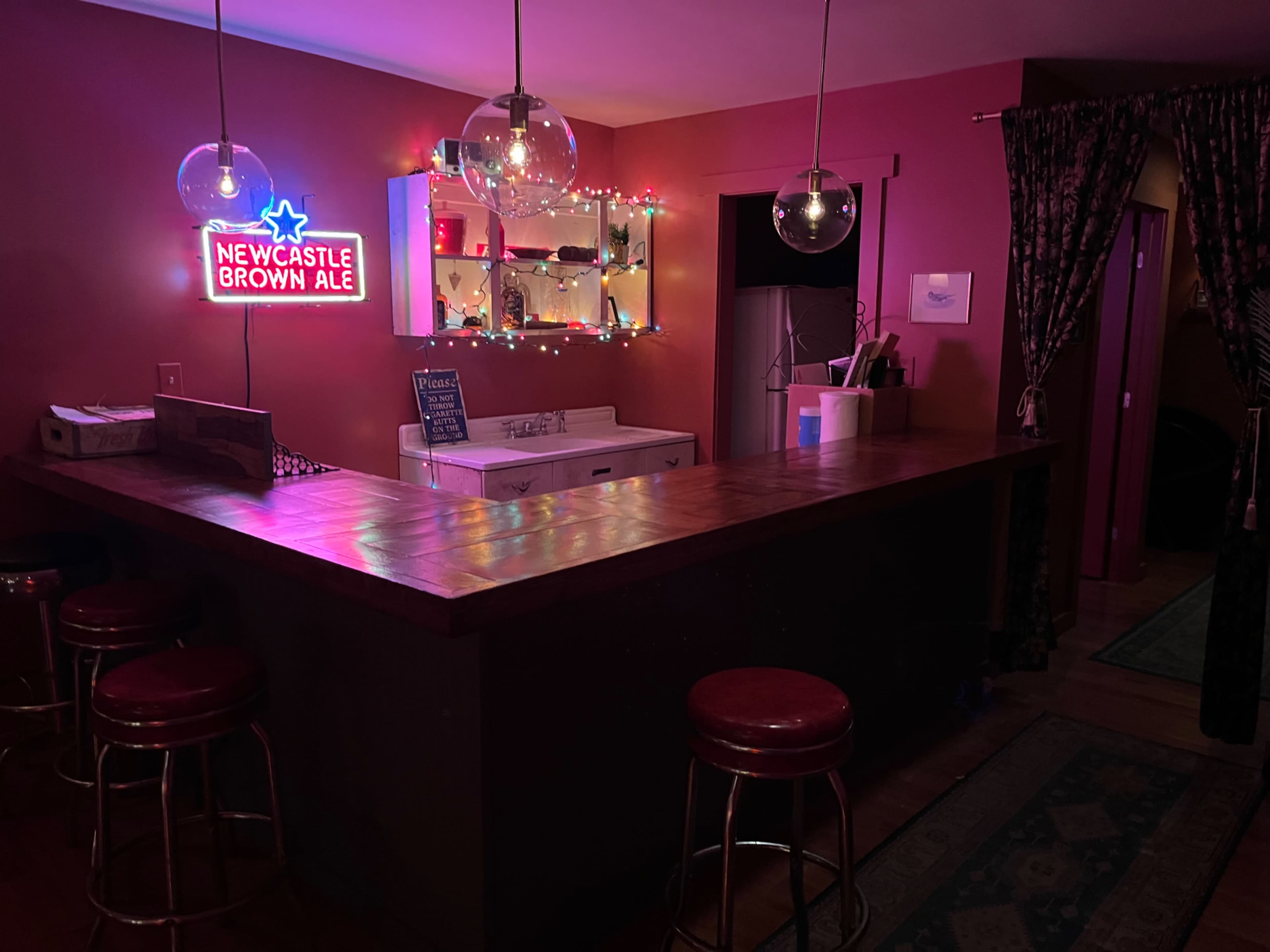 The scene depicts a dimly lit bar area with a wooden counter, red stools, a neon "Newcastle Brown Ale" sign, and colorful string lights in the background.