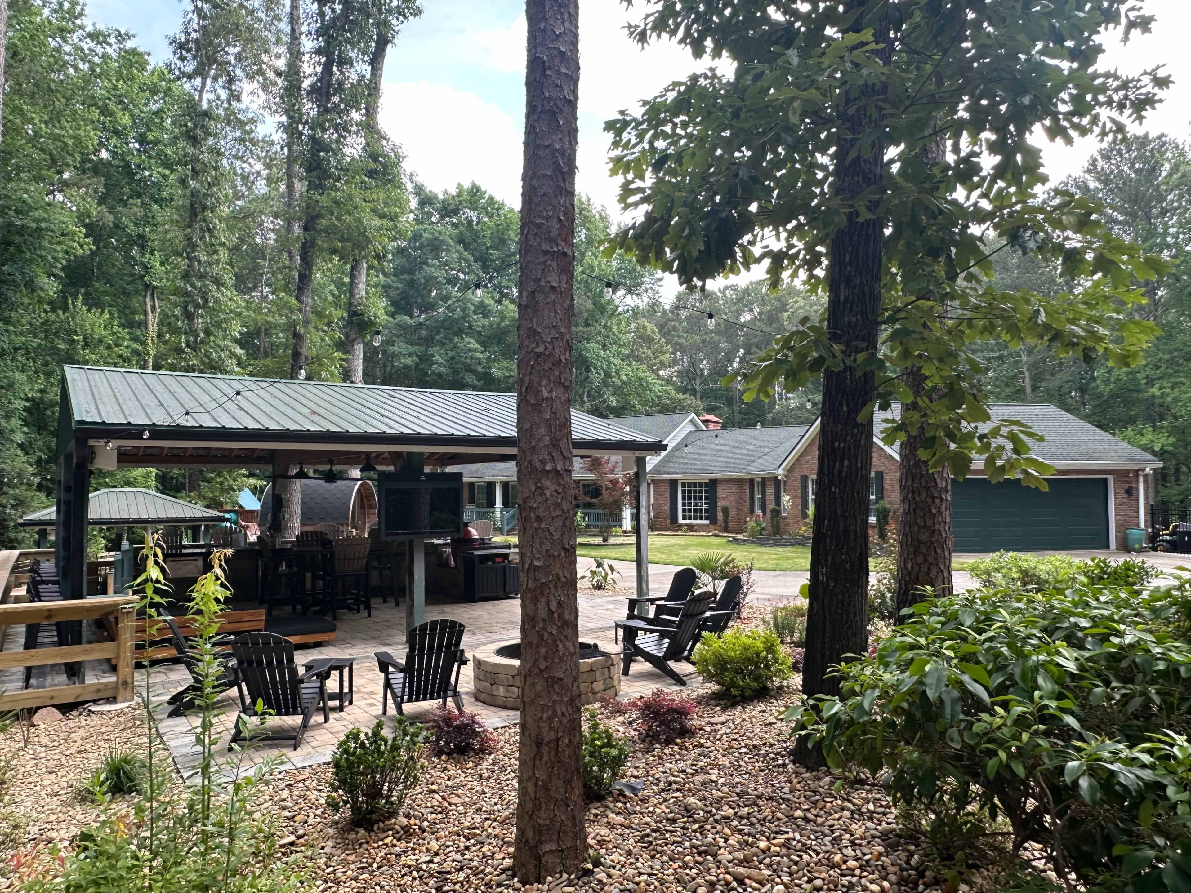 A patio area with outdoor seating and a barbecue setup is located beside a house surrounded by trees.