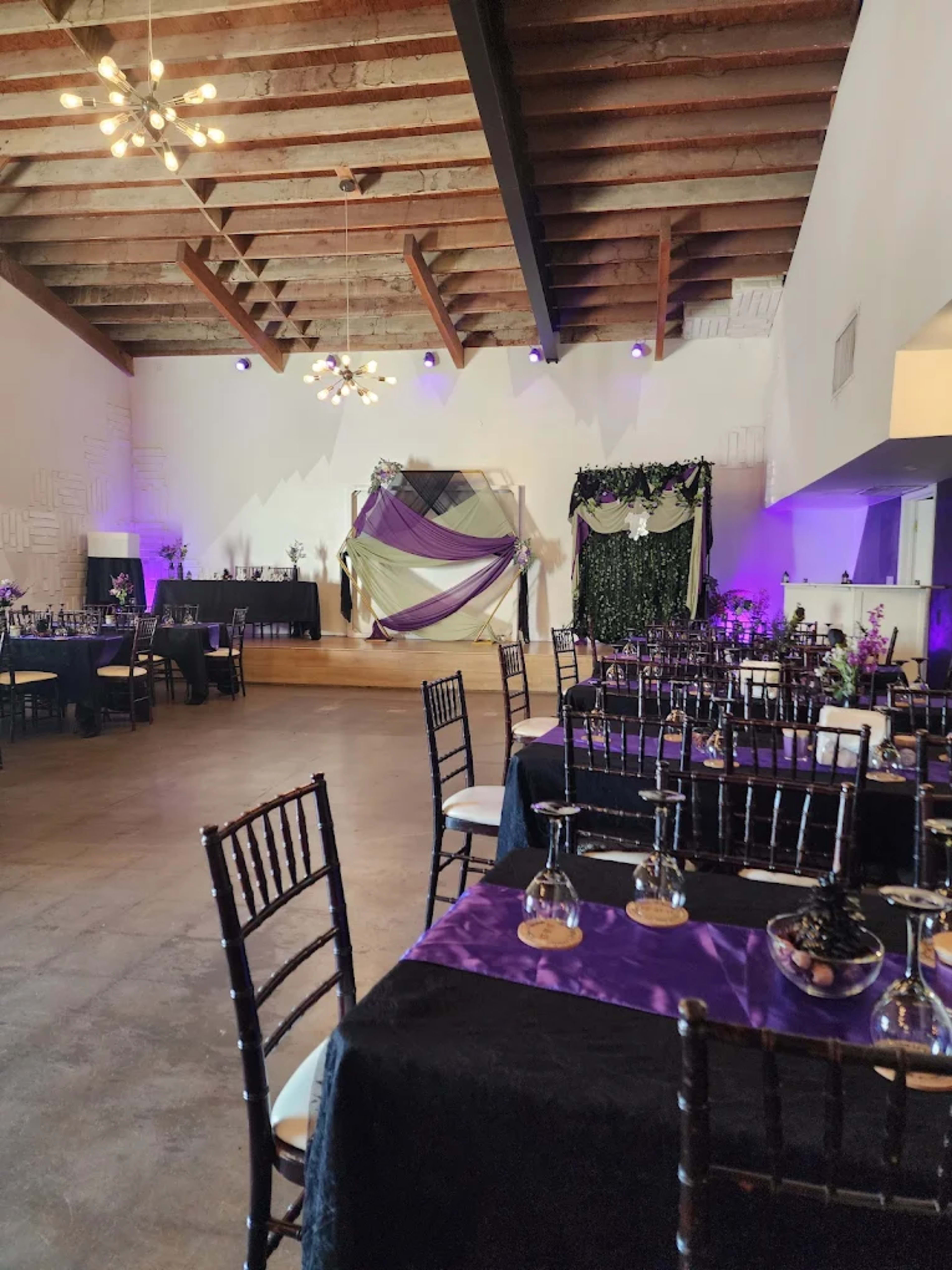 The image shows a spacious event hall decorated with purple and black table settings, featuring elegant chairs and decorative drapes along the walls.
