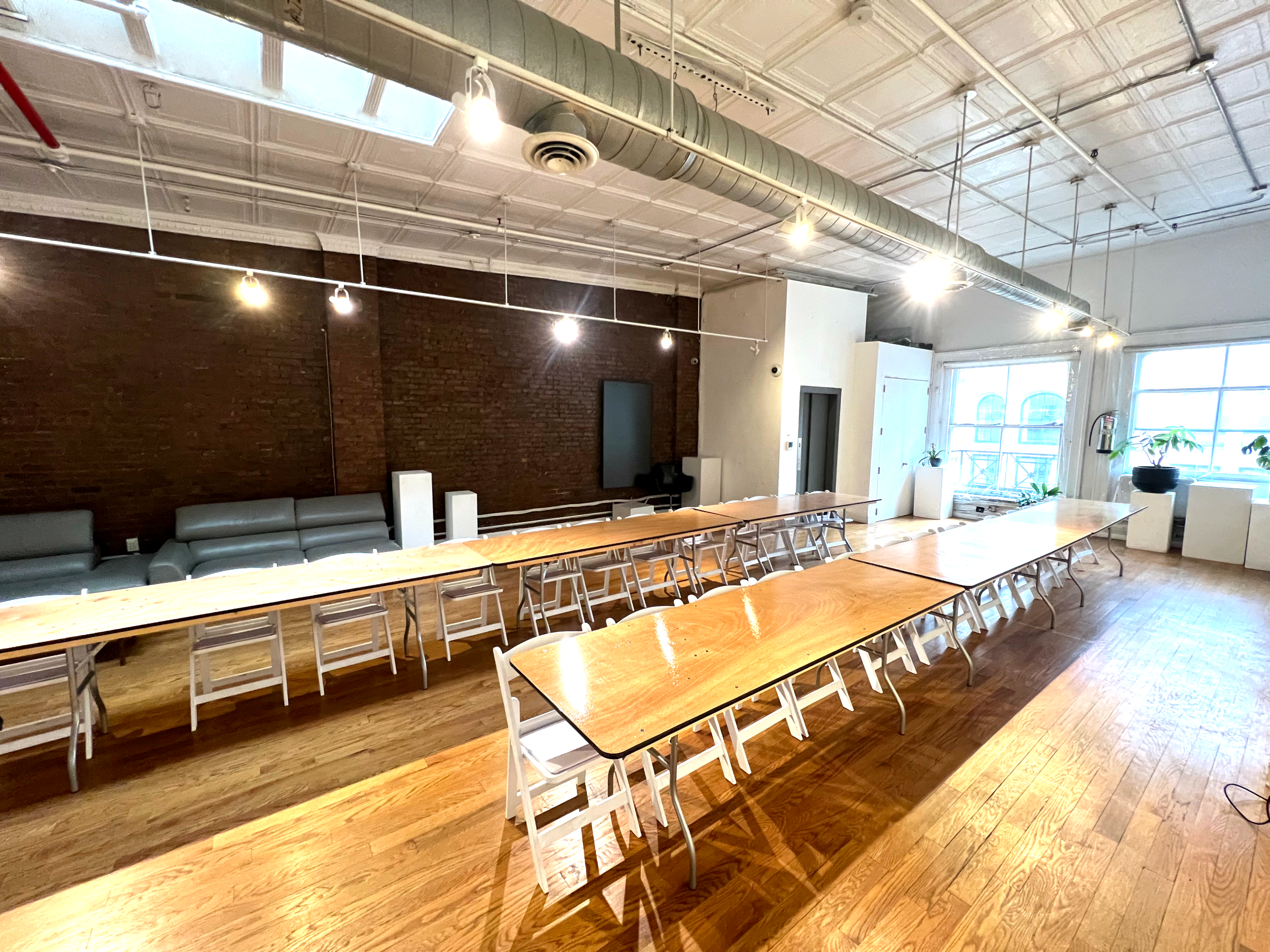 A spacious room features several long tables arranged for a gathering, with a backdrop of exposed brick walls and large windows.