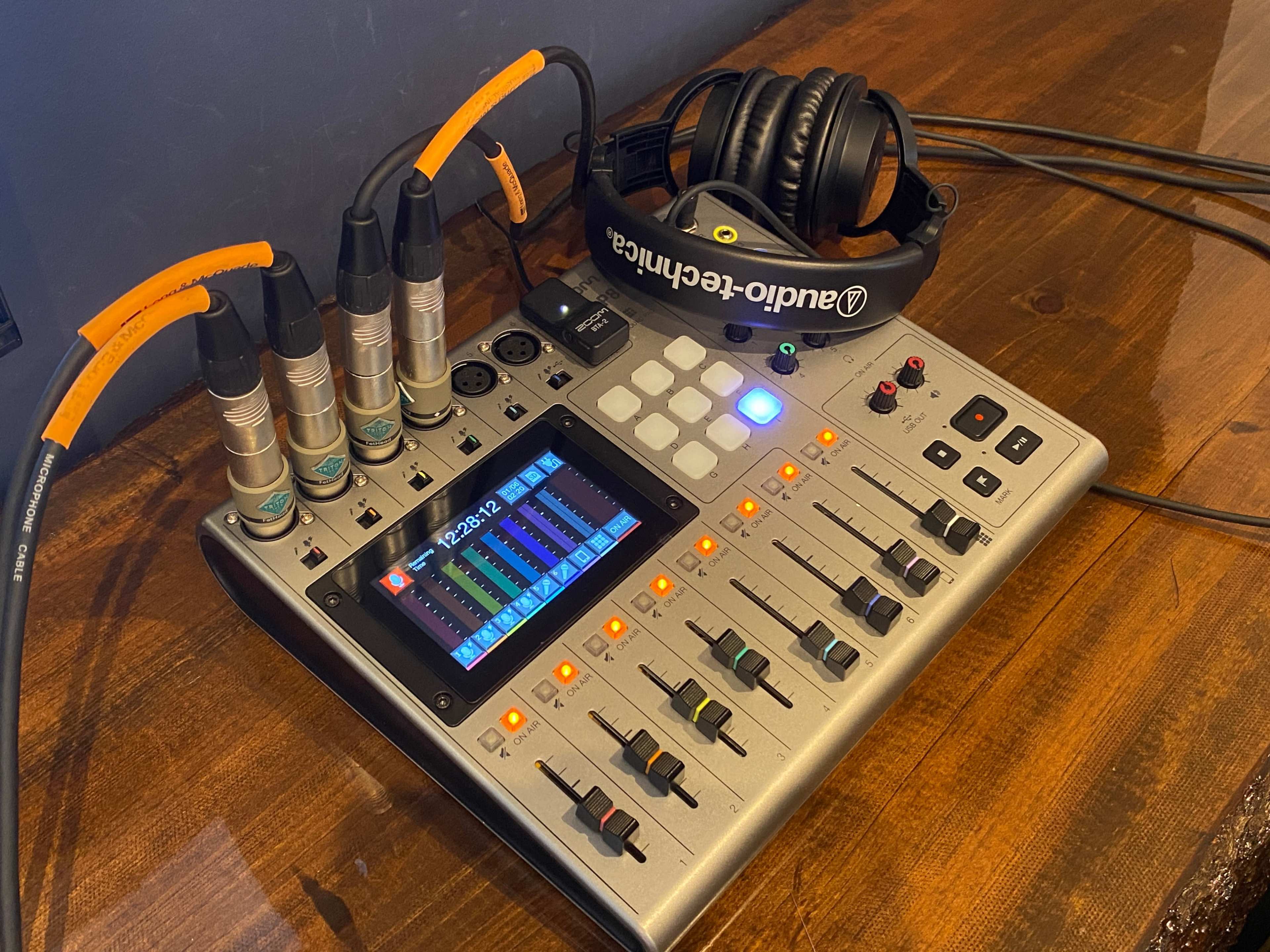 A digital audio mixer with multiple input jacks, faders, and a display is placed on a wooden table next to a pair of headphones.