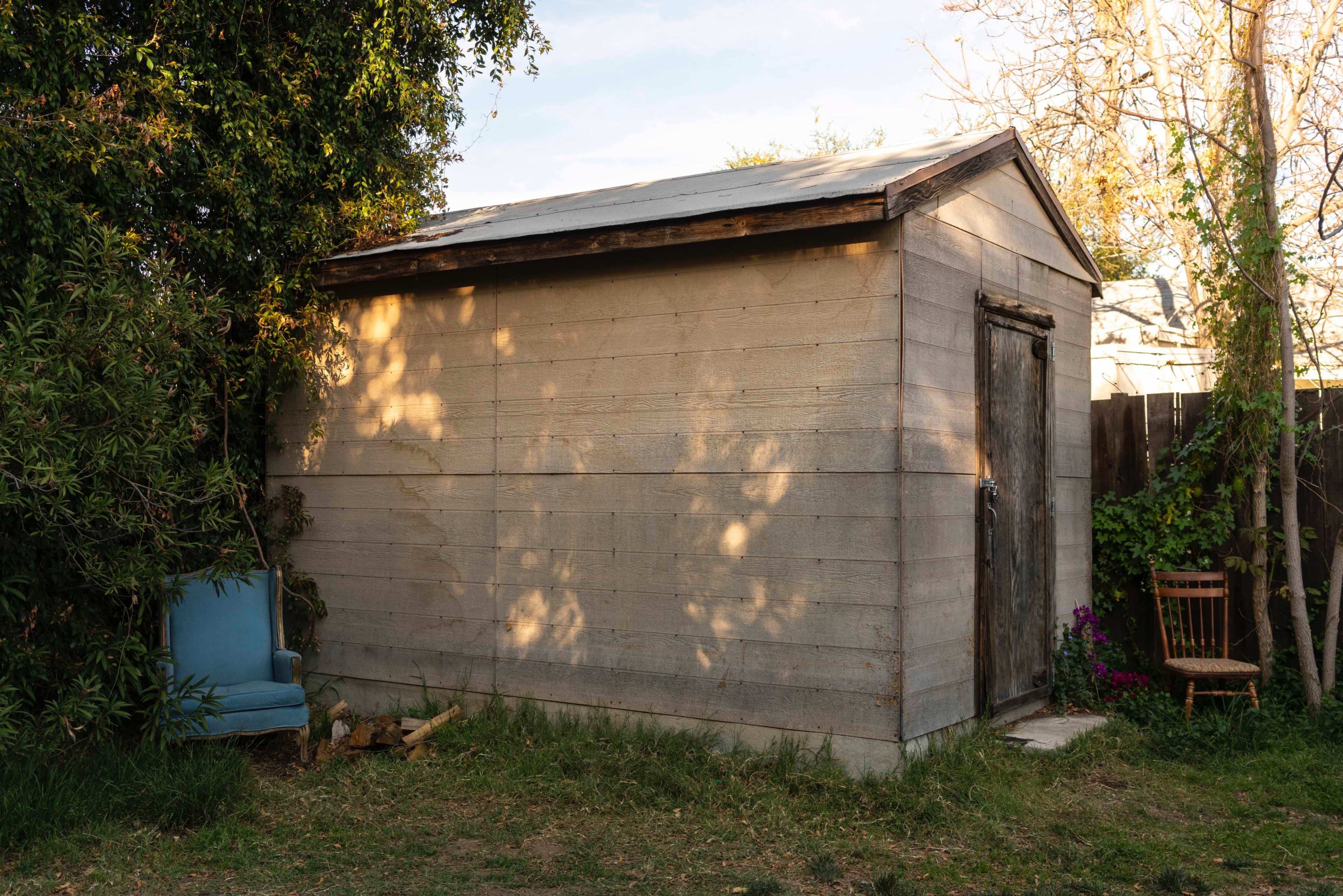A simple wooden shed with a metal roof sits in a grassy area, adjacent to a blue chair and a wooden chair.