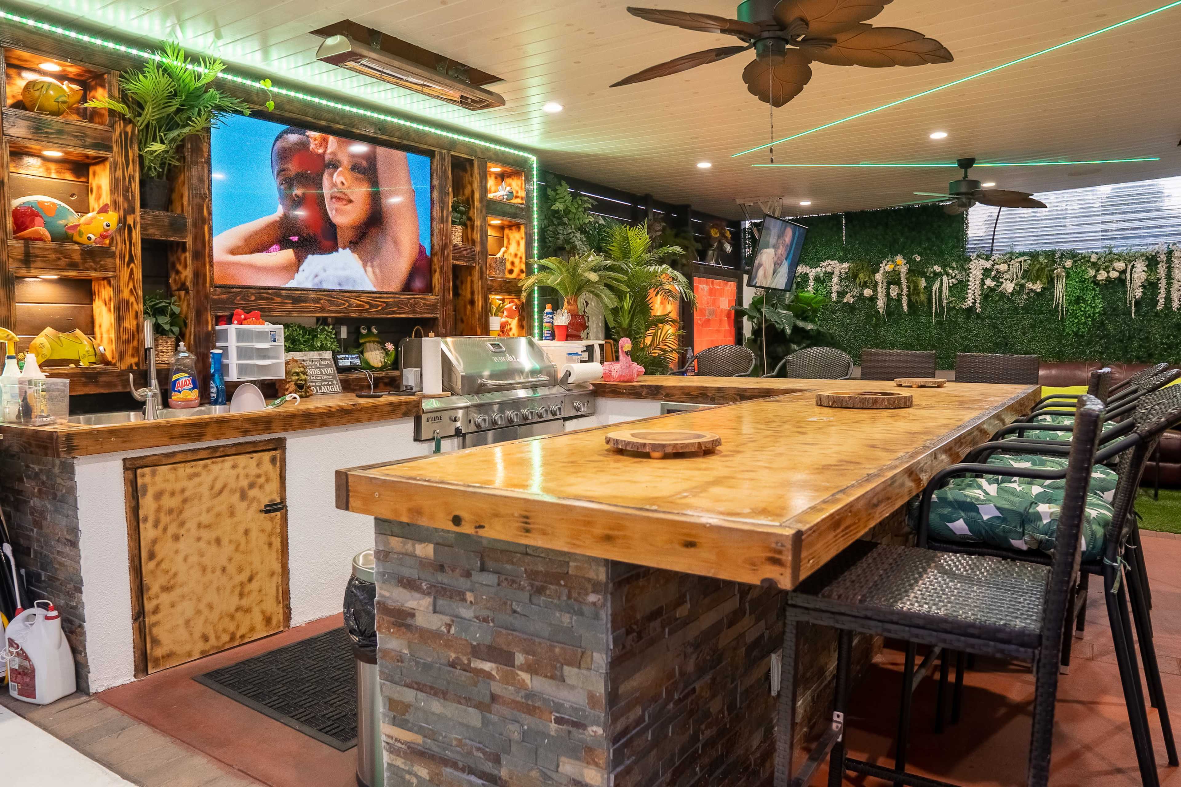 A well-decorated outdoor bar area featuring a wooden countertop, comfortable seating, and a large TV mounted on the wall.