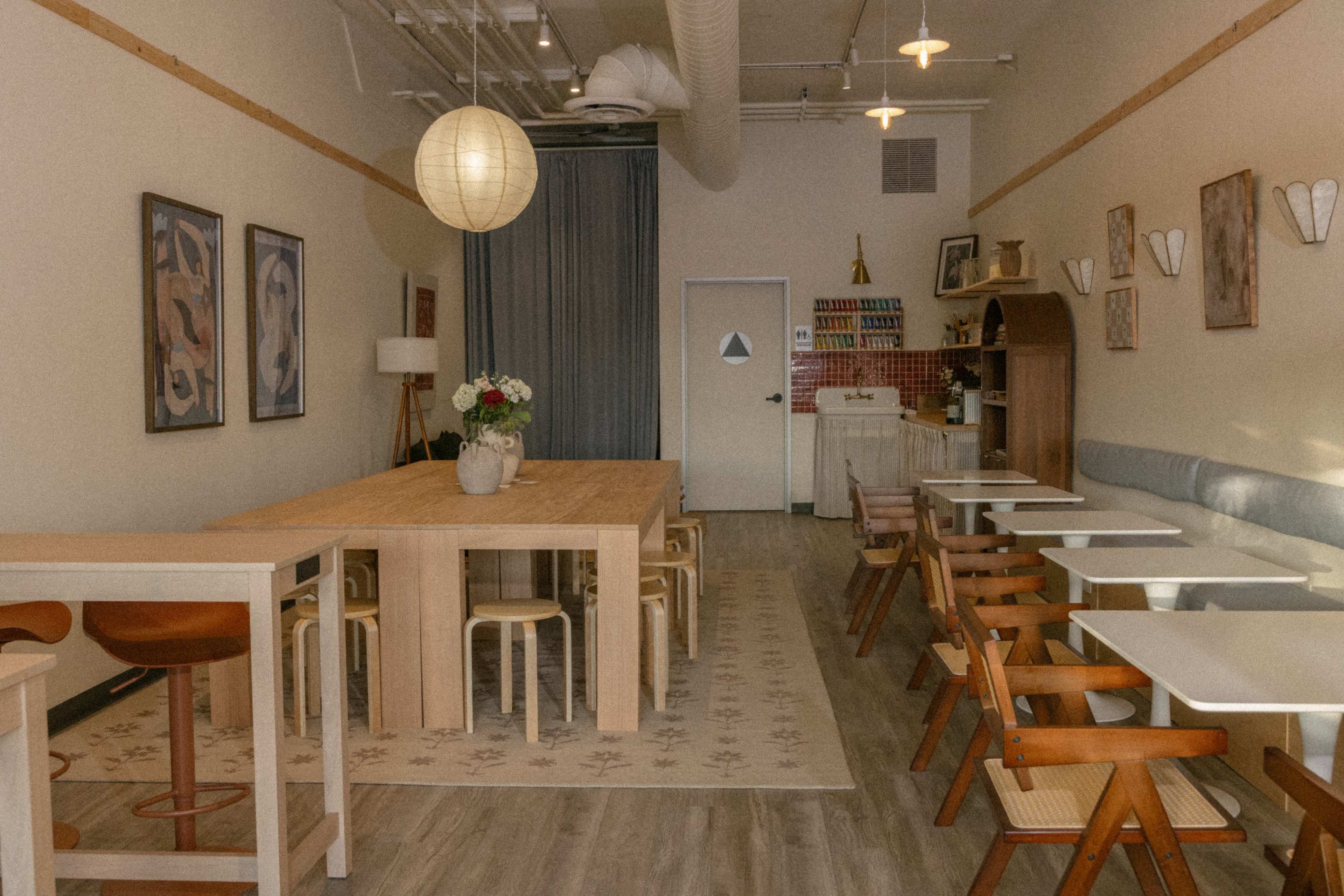 The image shows a light-colored café interior with wooden tables, chairs, and artwork on the walls.