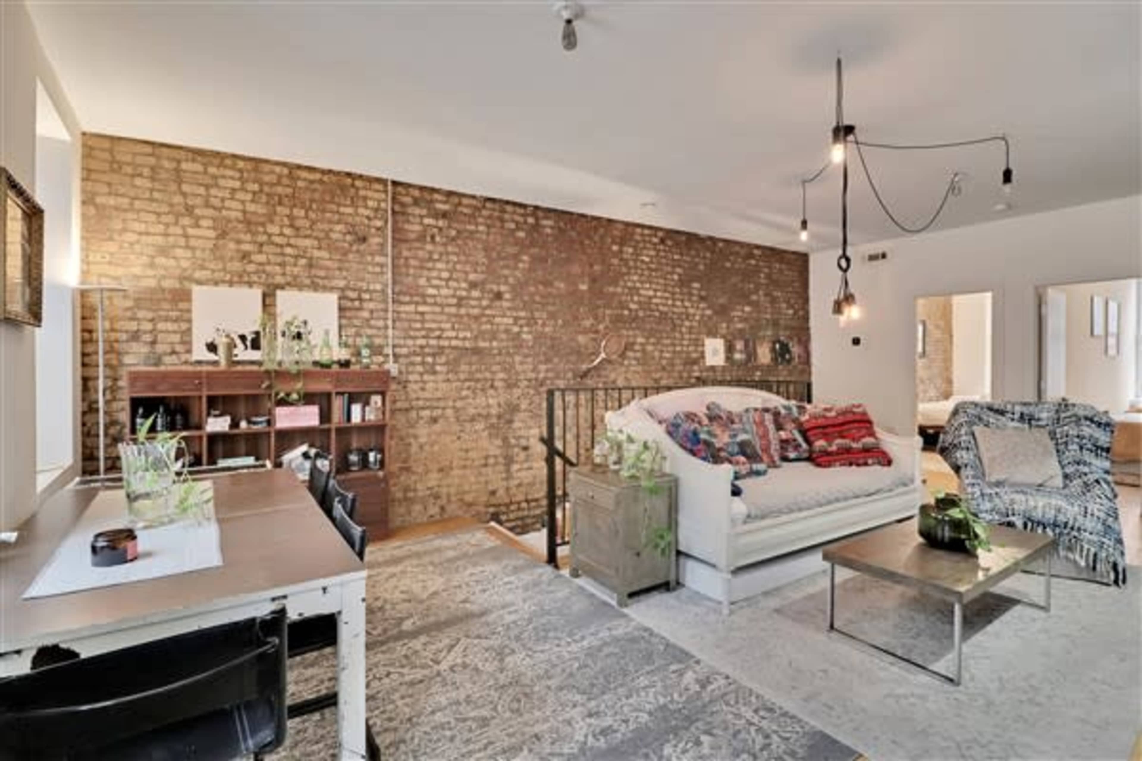 A living area with a brick wall, a white sofa adorned with colorful throw blankets, a wooden bookshelf, and a coffee table on a patterned rug.