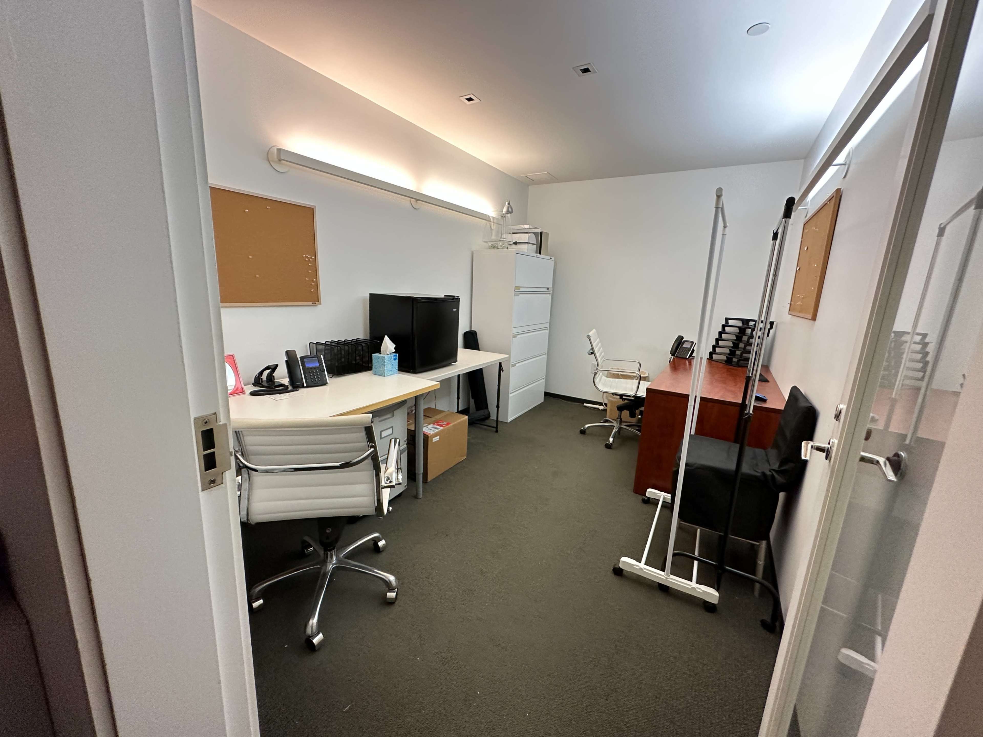 A small office space featuring two desks, office chairs, a large television, a filing cabinet, and a wall-mounted light.