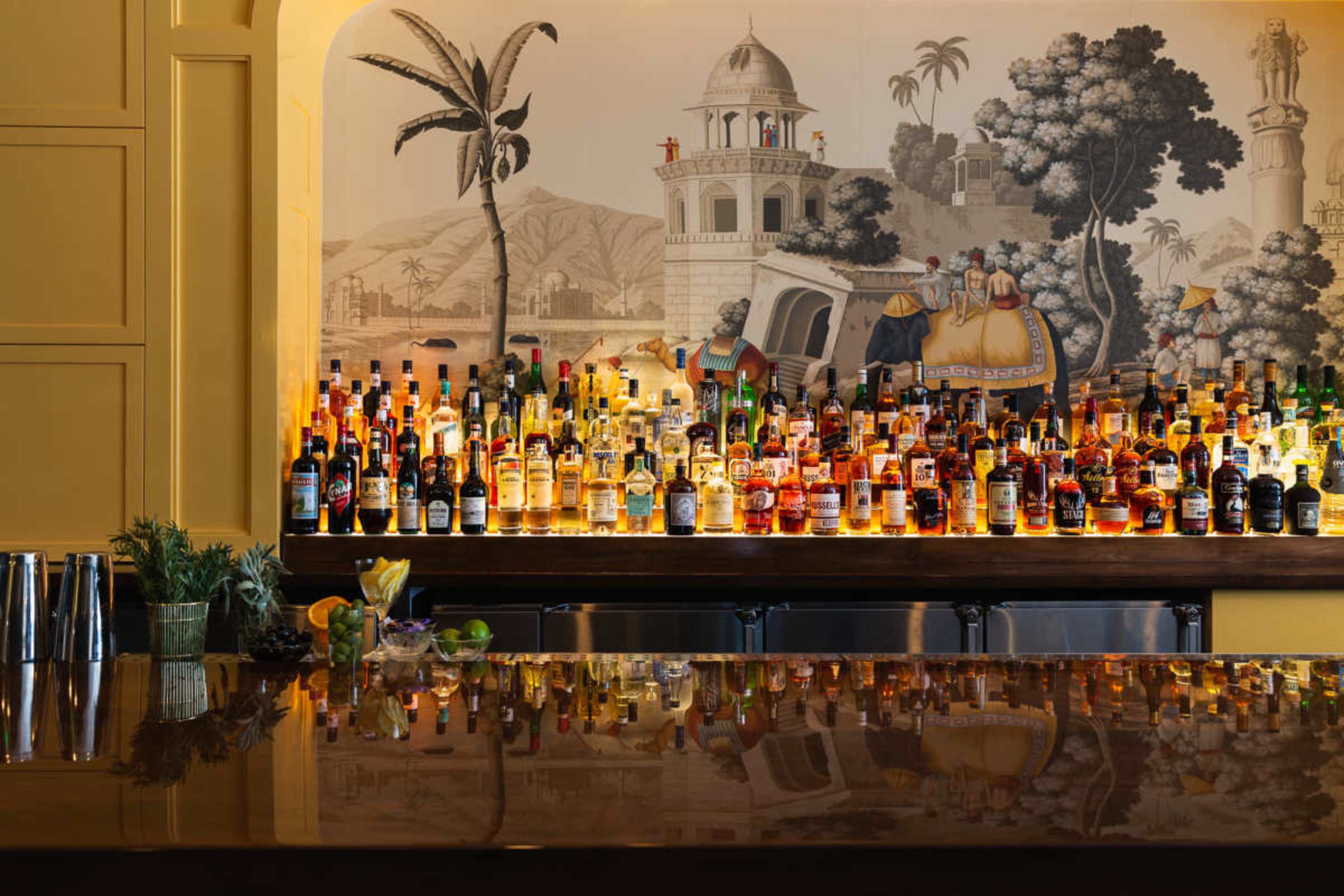 A well-stocked bar showcases a variety of colorful bottles against a detailed mural featuring tropical scenery and architecture.