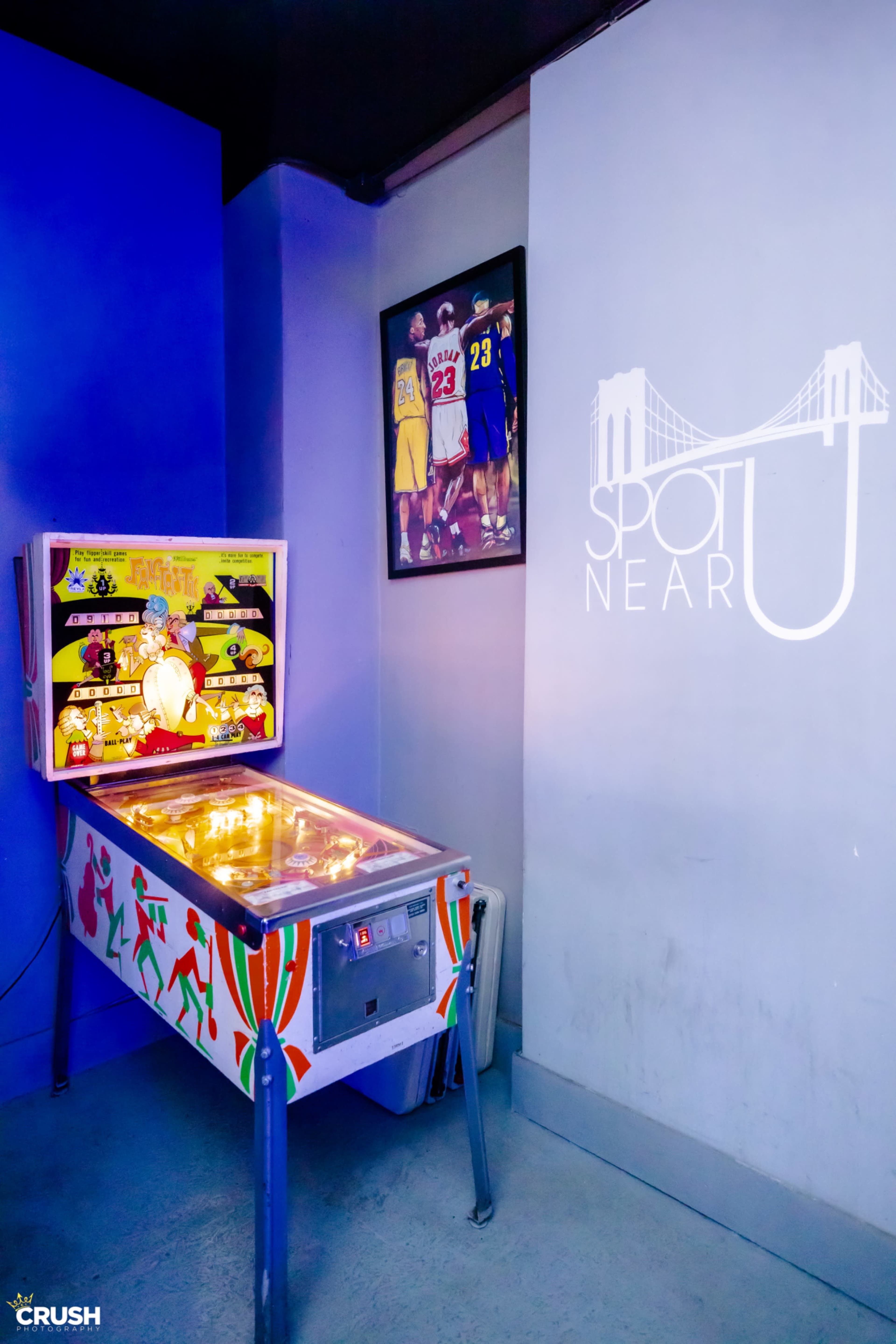 A pinball machine sits against a purple wall, next to a framed photo of basketball players, with the words "SPOT NEAR U" displayed on the wall.