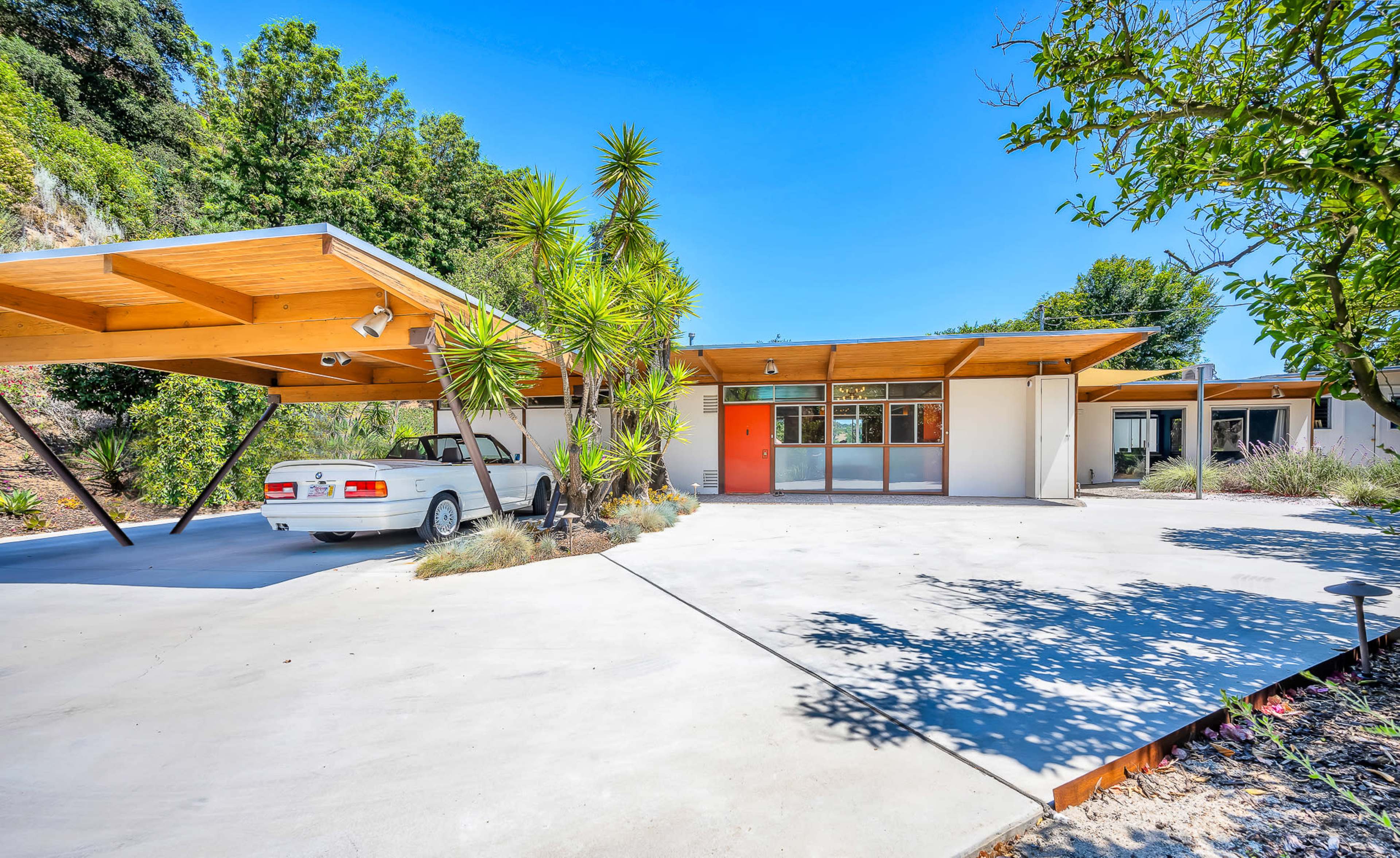 A modern single-story house features a carport with a parked white car and a vibrant orange door, surrounded by various plants and trees.