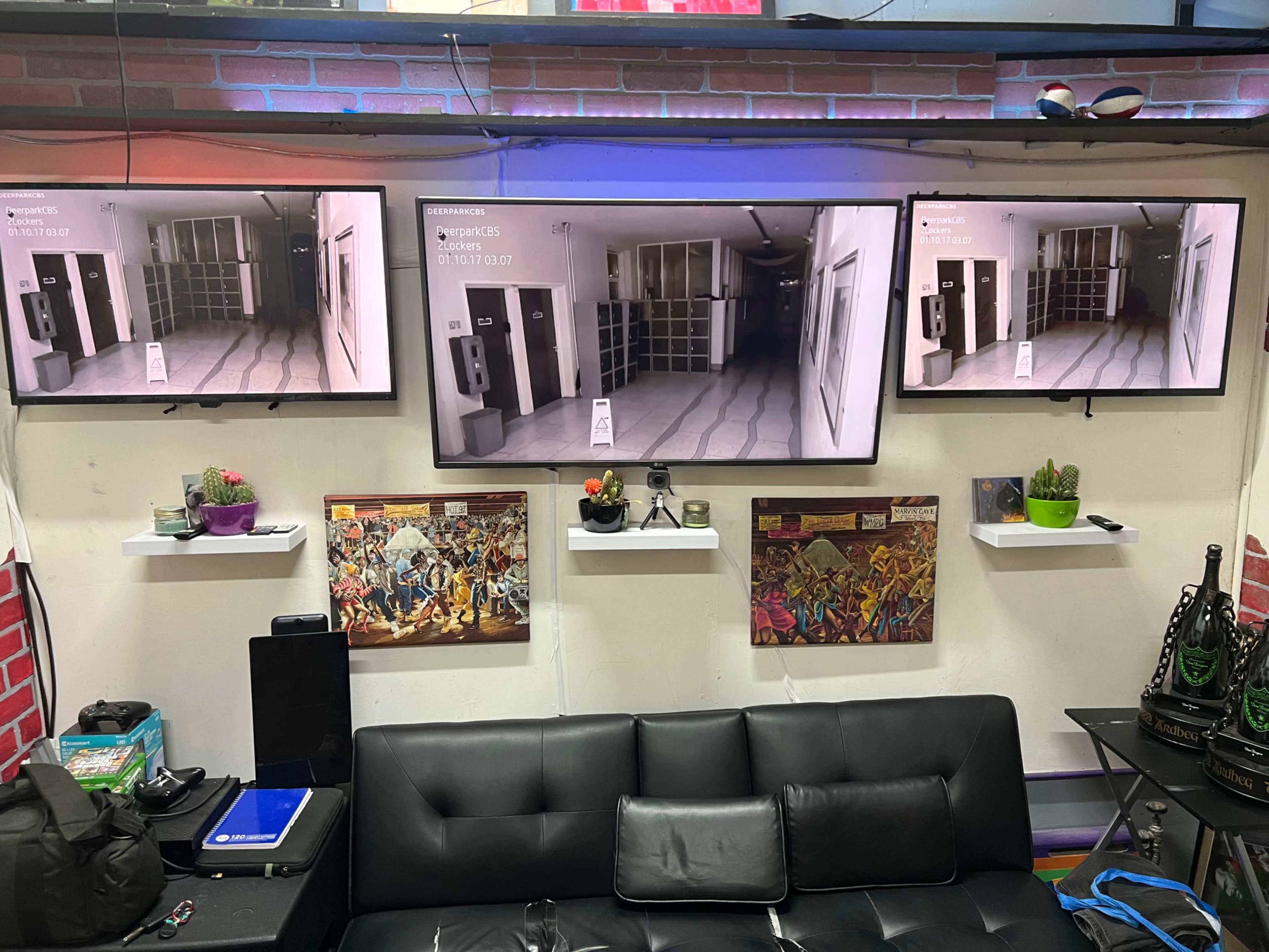 Three wall-mounted televisions display security footage, while plants and artwork are arranged on shelves below in a room with a black couch.