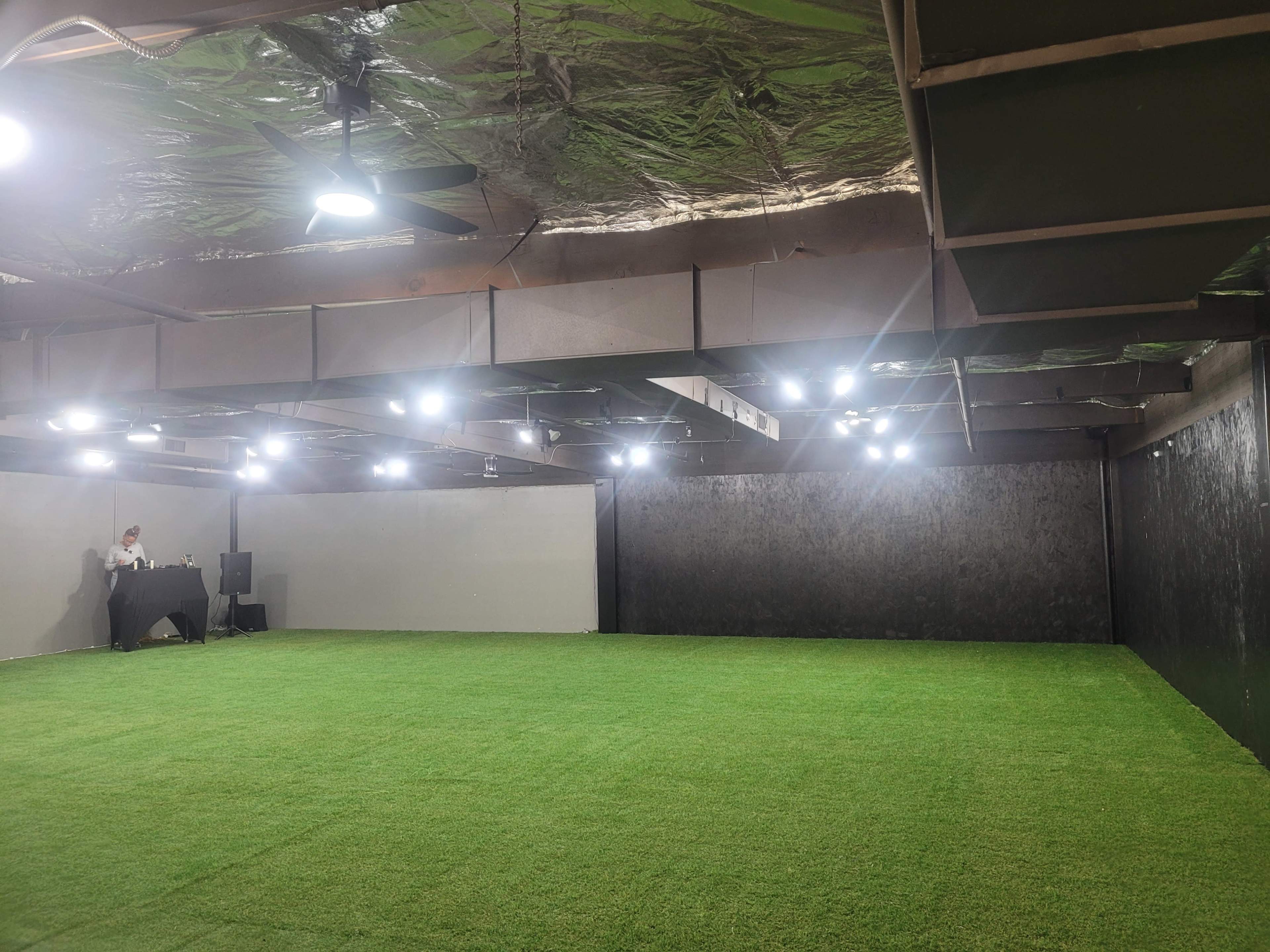 The image shows a spacious interior room with artificial grass flooring and spotlights, featuring a black wall and a small setup with a table and equipment on one side.
