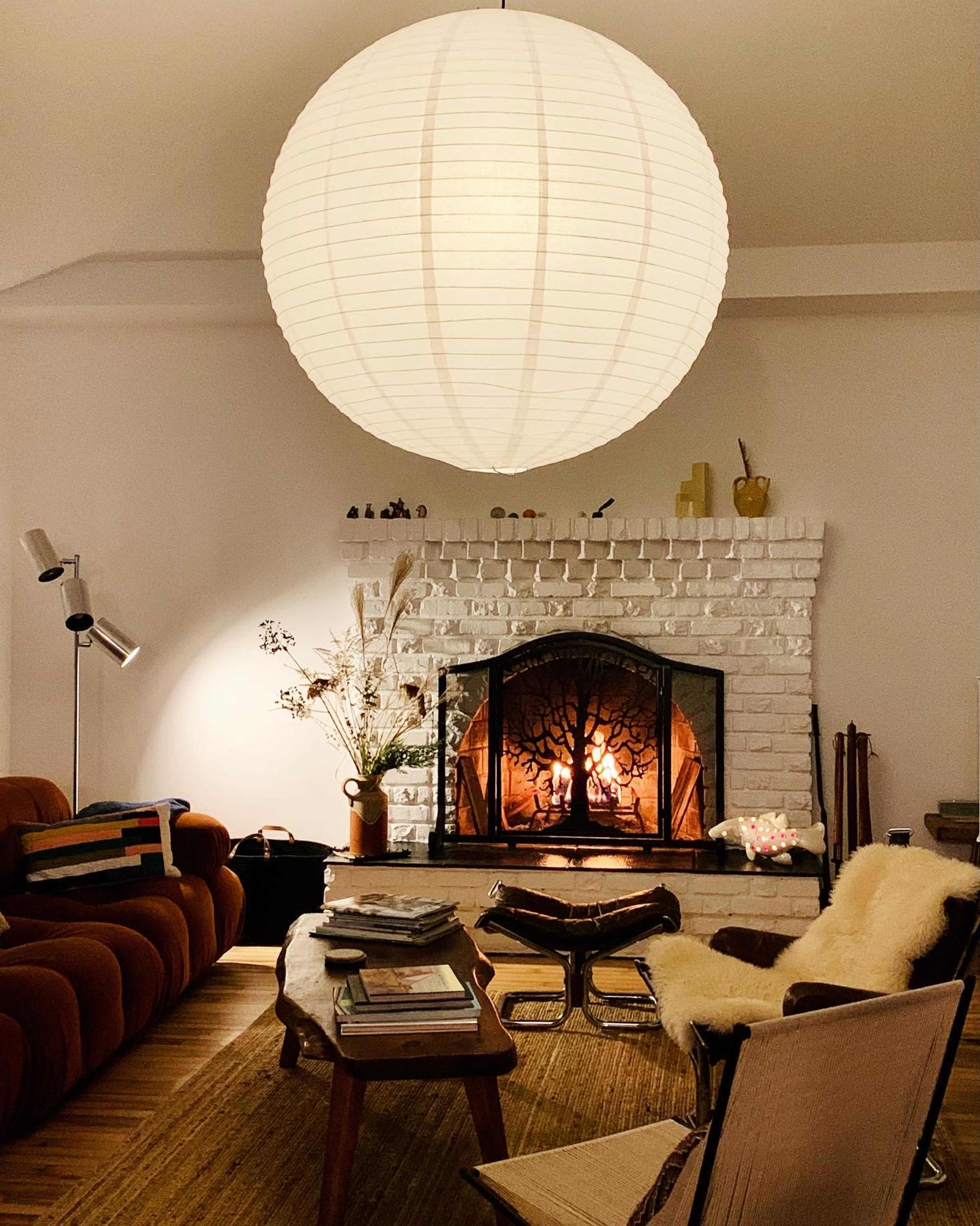 A warm-lit living room features a large paper lantern hanging above a stone fireplace with a fire burning, surrounded by comfortable furniture and decorative items.