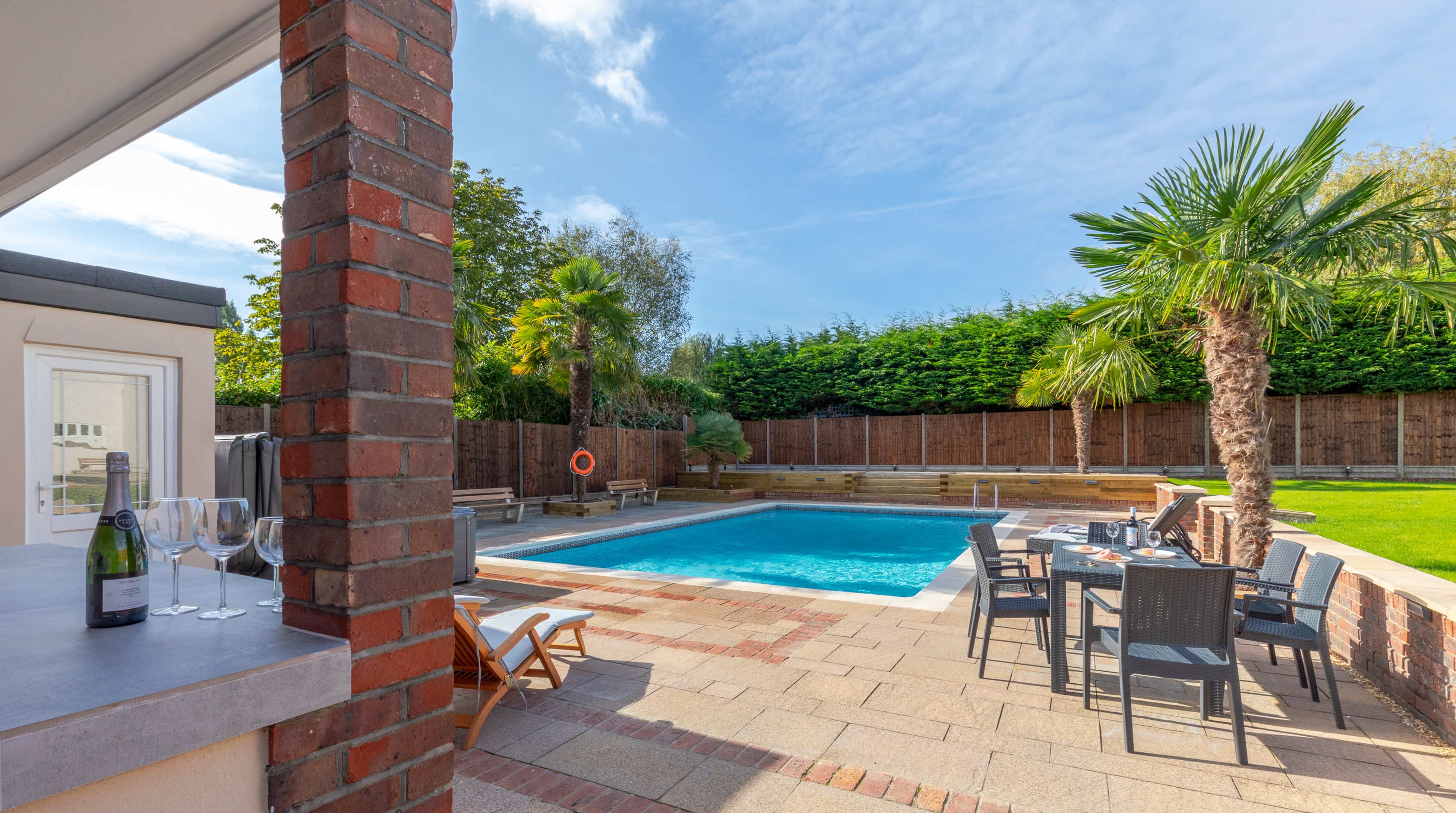 A backyard pool area with a swimming pool, lounge chairs, a dining table, and palm trees surrounded by greenery and wooden fencing.