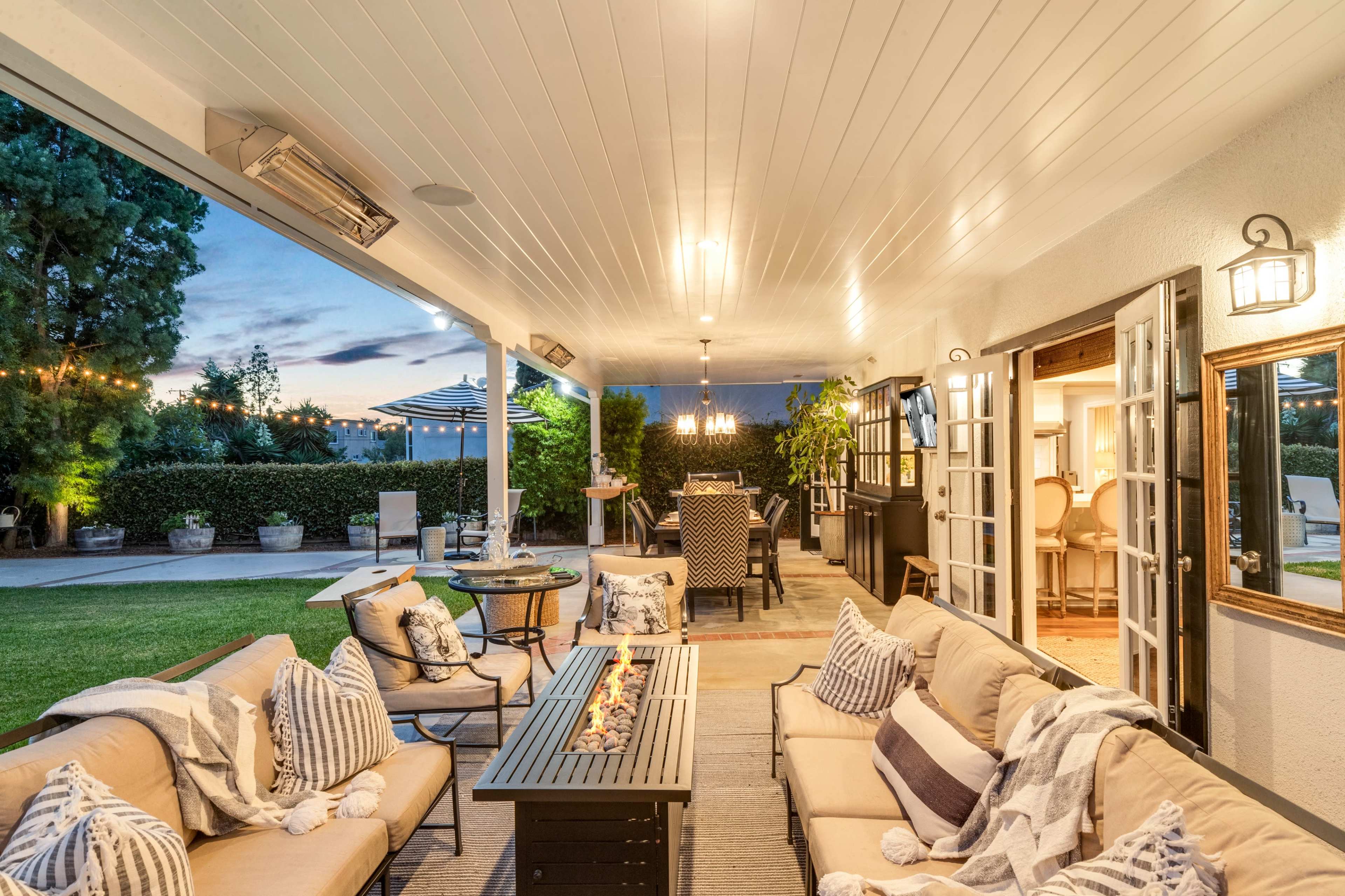 A covered outdoor patio featuring a seating area with a fire table, potted plants, and a dining space visible in the background.